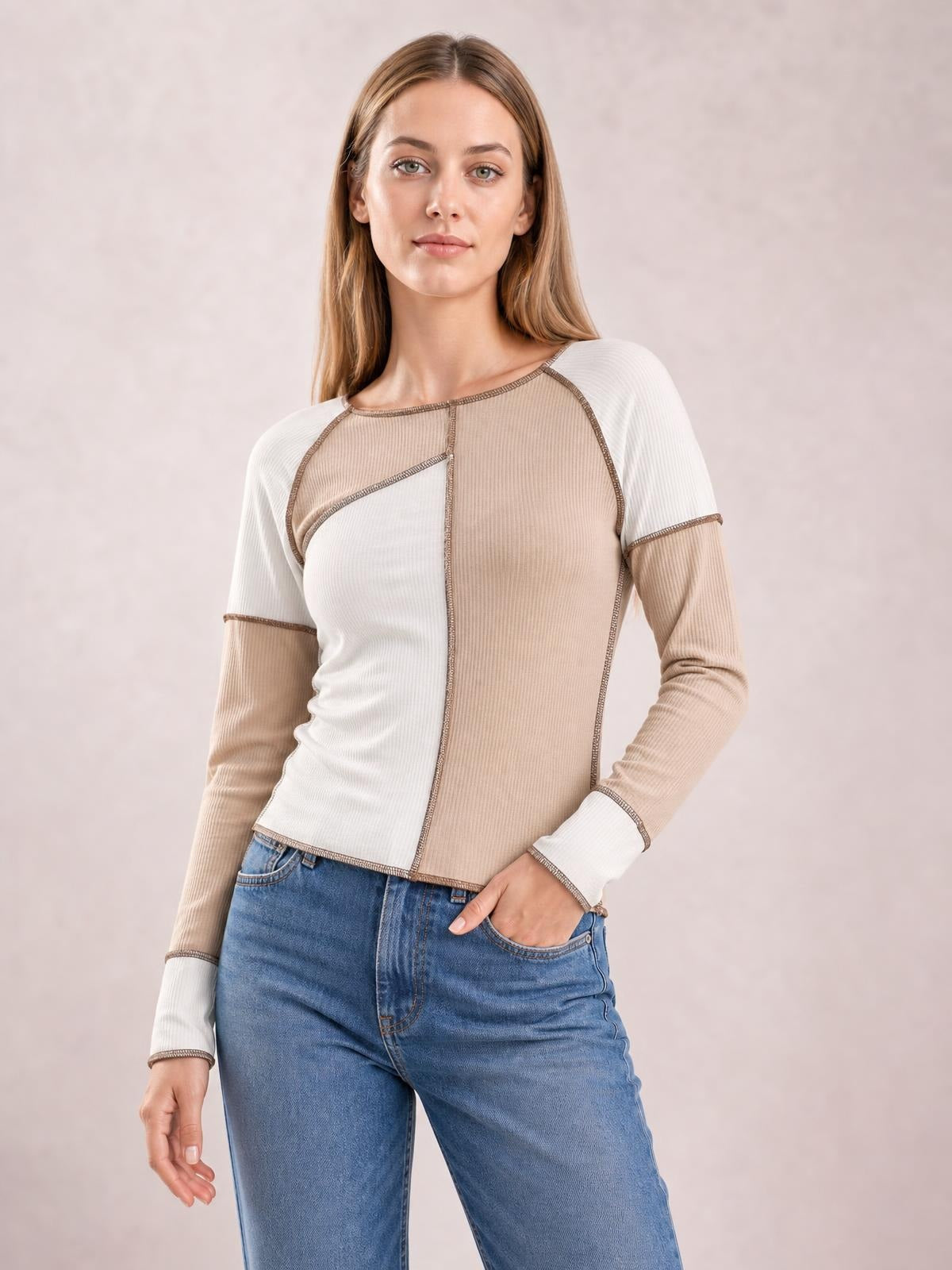Model poses with one hand in her jeans pocket, wearing a fitted taupe and white color block top with ribbed long sleeves, a crew neck, and exposed seams.