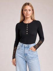 A model stands in a black button-front top with a round neckline, fitted long sleeves, and crochet lace hem cuffs, paired with light-wash jeans.