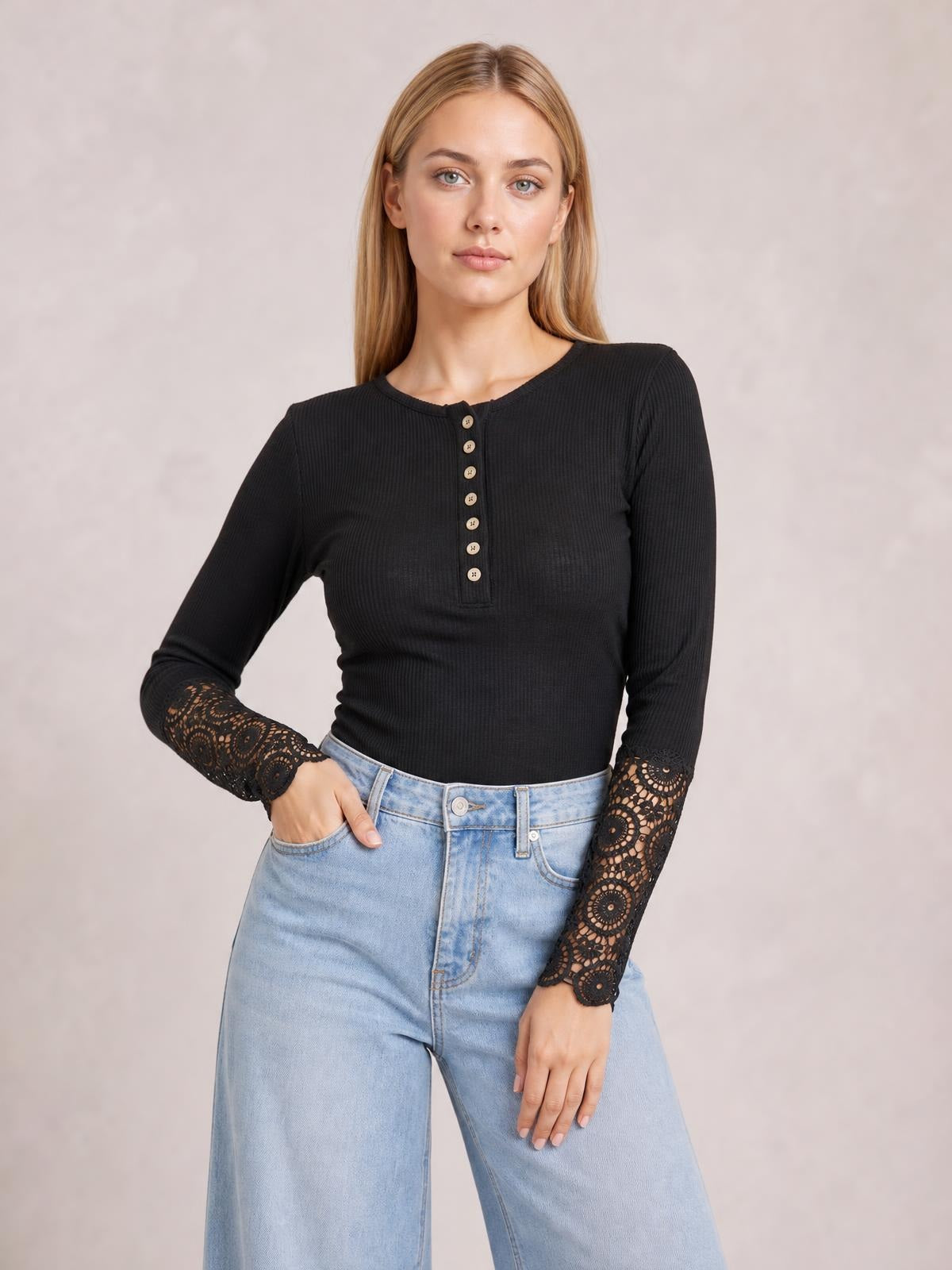 Model poses with one hand in her jeans pocket wearing a black ribbed button-front top with long crochet lace hem sleeves and a fitted silhouette.