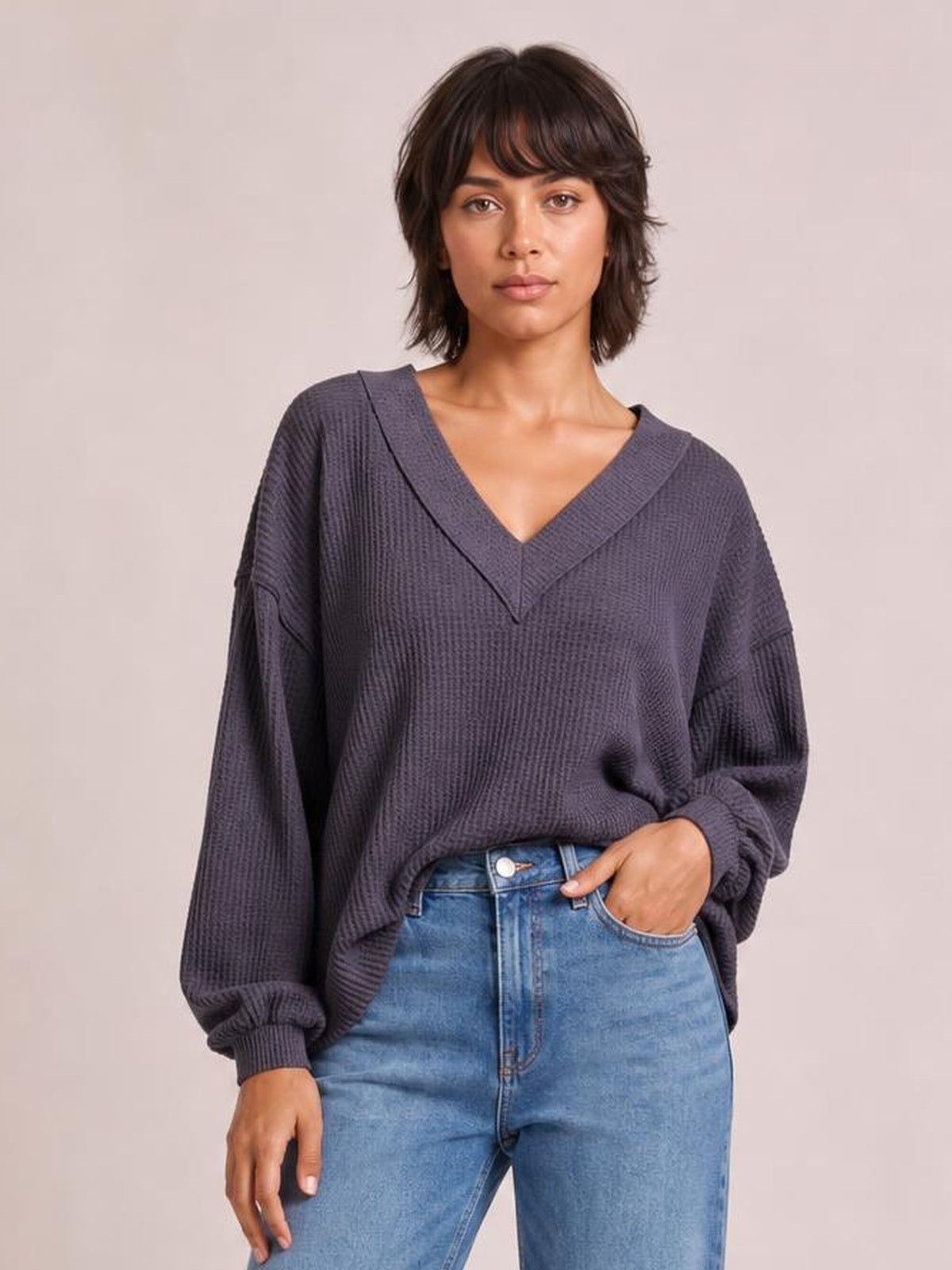 Model wearing a charcoal ribbed textured V-neck long sleeve T-shirt with exposed seams, relaxed silhouette, and ribbed cuffs, posed with one hand in her jeans pocket.