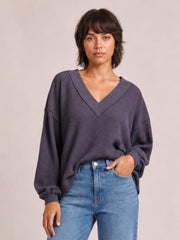 Model wearing a charcoal ribbed textured V-neck long sleeve T-shirt with exposed seams, relaxed silhouette, and ribbed cuffs, posed with one hand in her jeans pocket.