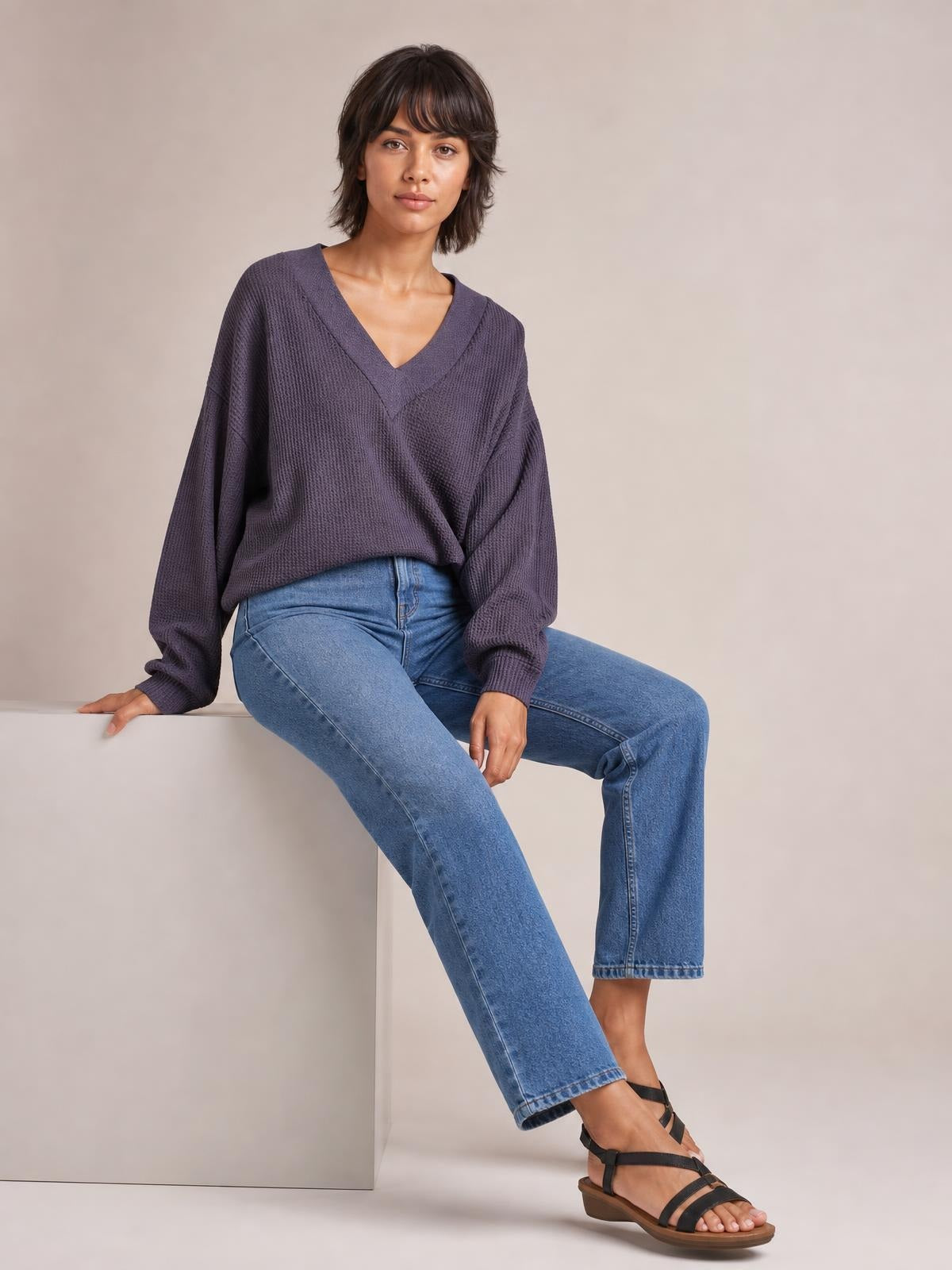 A seated model wears a deep plum ribbed textured V-neck long sleeve T-shirt with exposed seams and a relaxed, slightly loose silhouette.