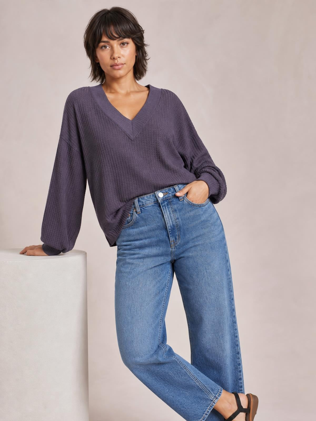 A model leans against a block wearing a plum ribbed textured V-neck long sleeve T-shirt with exposed seams and a relaxed silhouette.