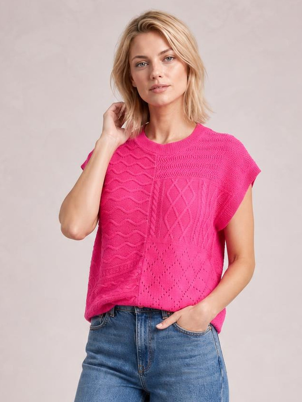 A model wears a bright pink cable-knit round neck cap sleeve sweater with a relaxed silhouette and mixed textured knit panels, touching her hair.