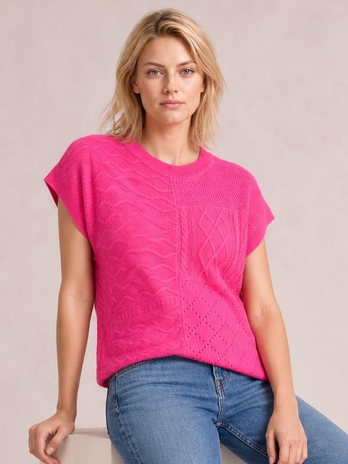 A seated model wears a bright pink cable-knit sweater with a round neck, cap sleeves, textured panel construction, and a relaxed straight silhouette.