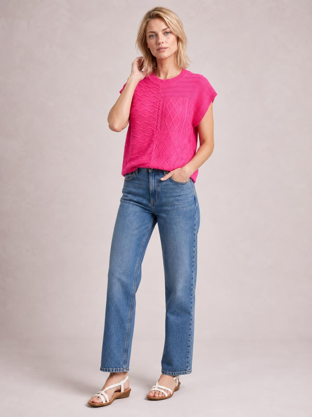 Model poses in a bright pink cable-knit round neck cap sleeve sweater with a relaxed straight silhouette and textured panel construction.