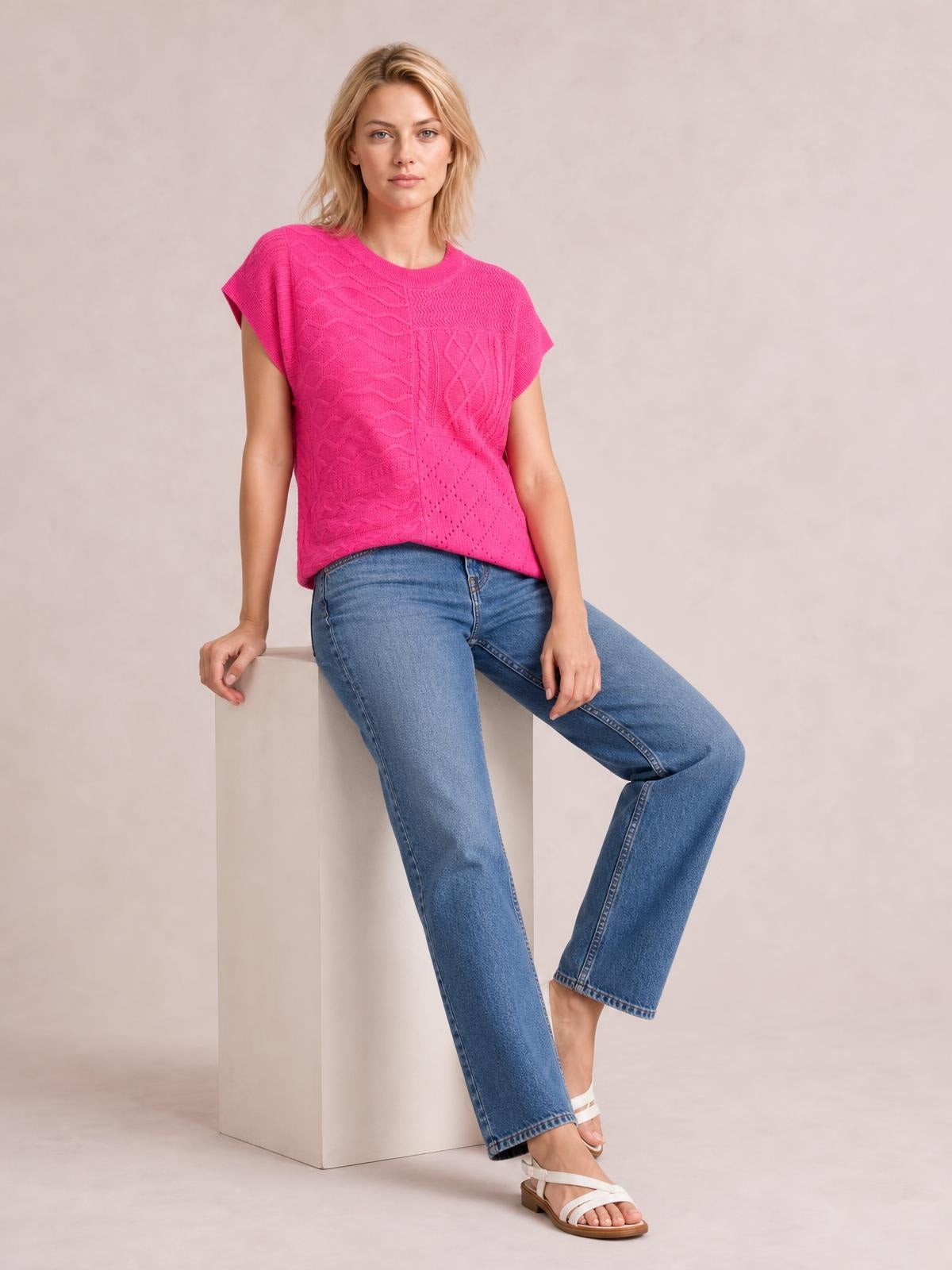 A model sits in blue jeans wearing a bright pink cable-knit round neck cap sleeve sweater with a relaxed, textured silhouette.