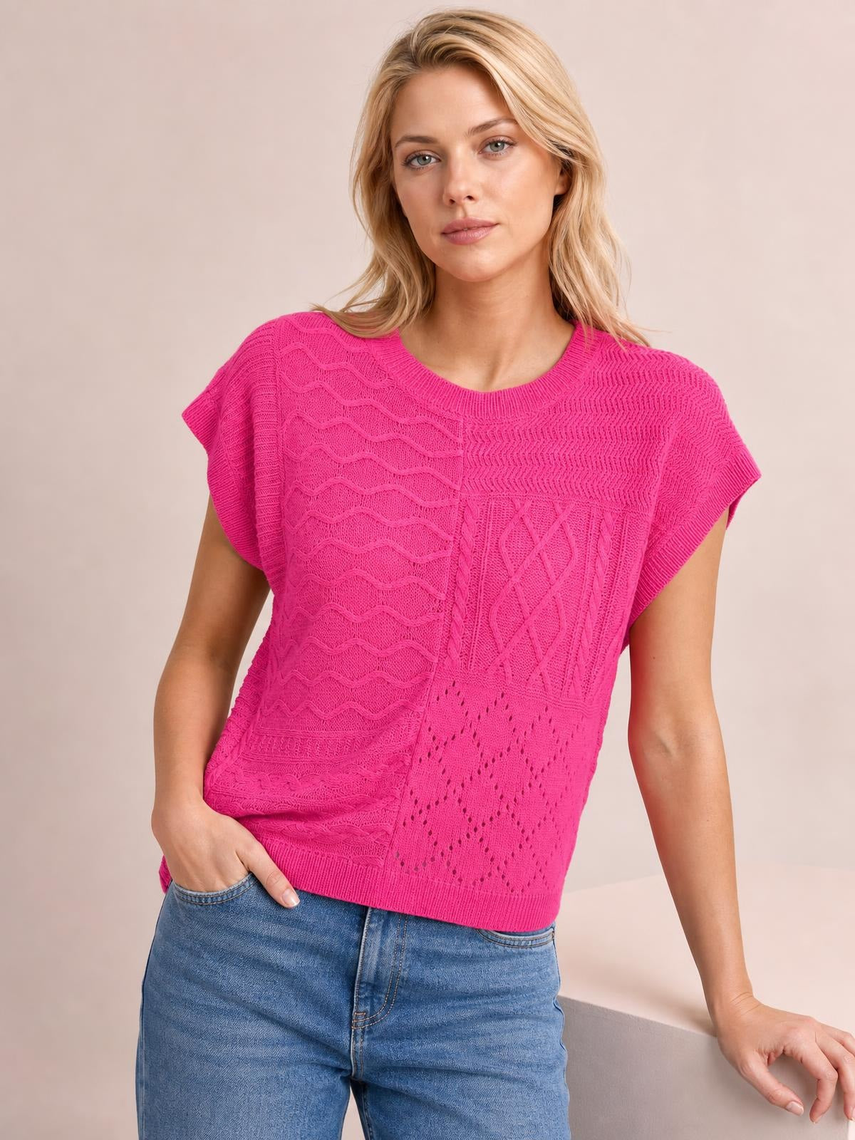 Model wears a bright pink cable-knit round neck cap sleeve sweater with textured panels, a relaxed silhouette, and a ribbed hem while posing with one hand in her jeans pocket.