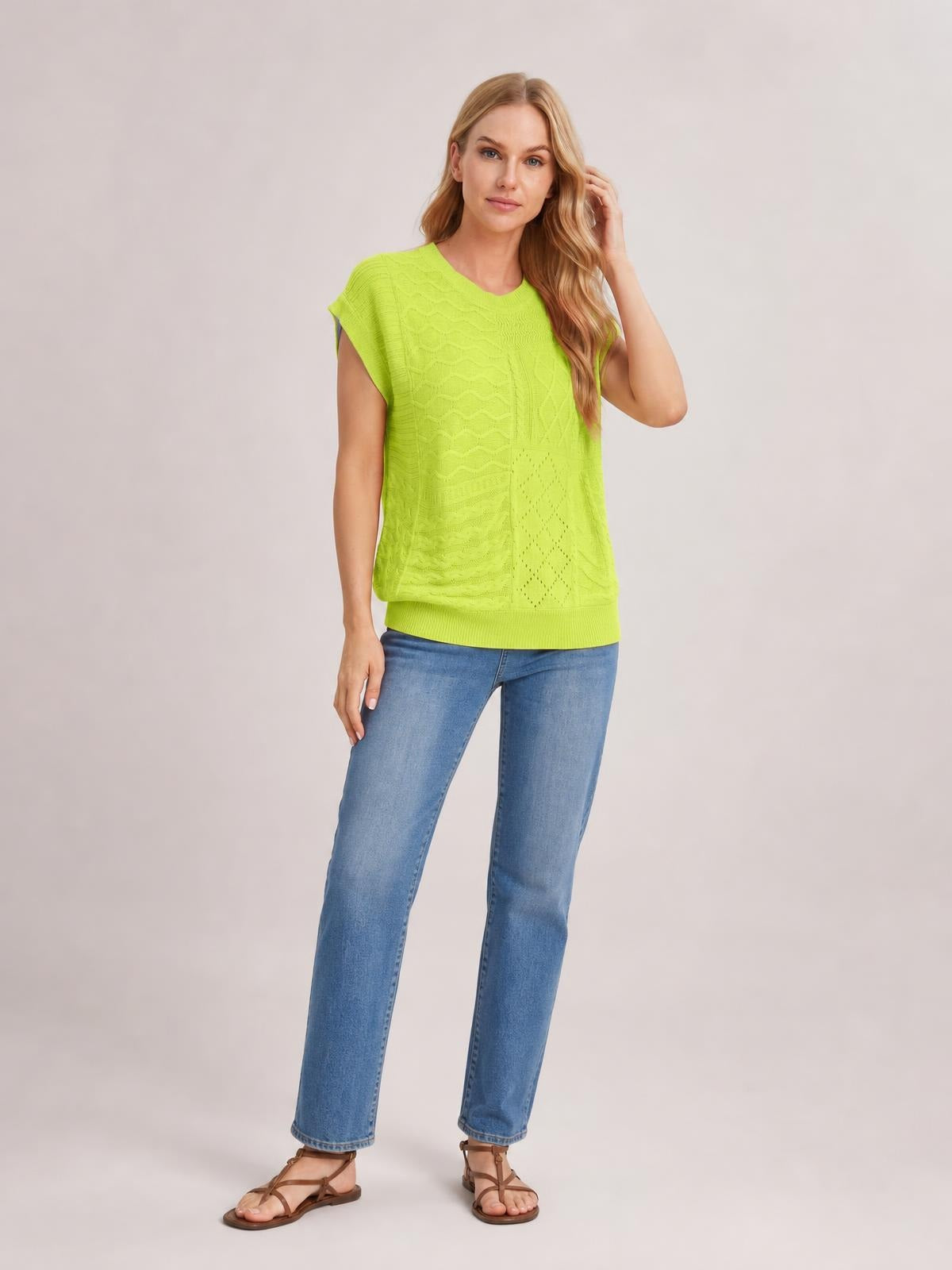 Model wearing a bright lime green cable-knit round neck cap sleeve sweater with a relaxed straight silhouette and ribbed hem, posing with one hand in her hair.