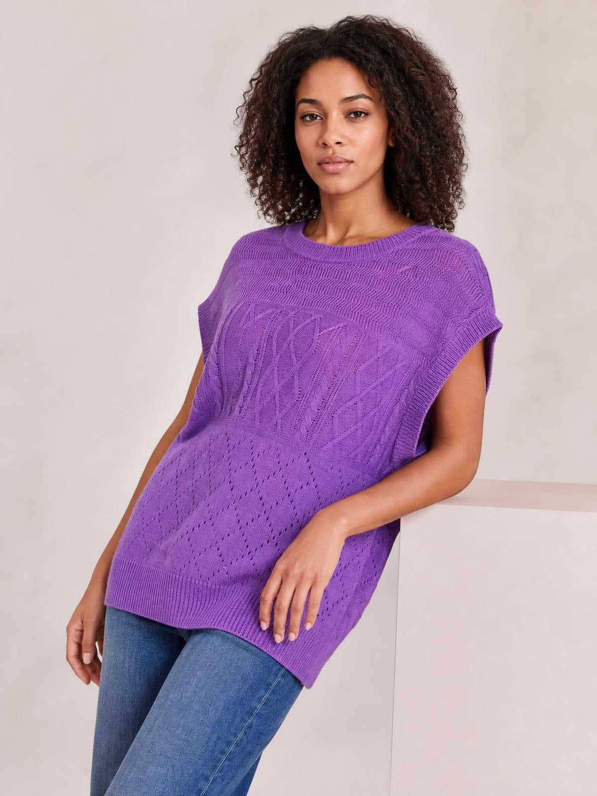 A model leans in a purple cable-knit sweater with a round neck, cap sleeves, relaxed silhouette, mixed knit textures, and a ribbed hem.