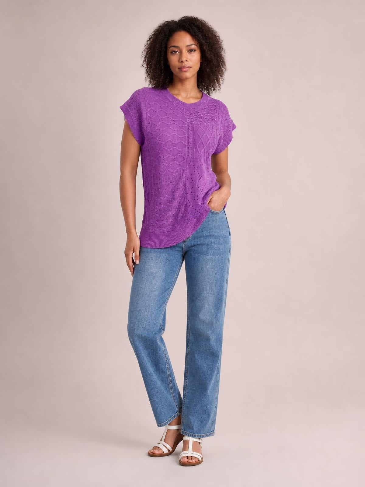 Model wears a purple cable-knit round-neck cap-sleeve sweater with a relaxed silhouette, ribbed trim, and textured panel construction, posing with one hand in her pocket.