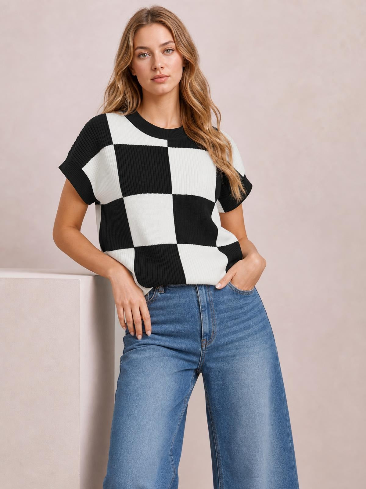 A woman wears a black-and-white checkered round-neck short-sleeve sweater with ribbed knit texture and a relaxed silhouette, posing with hands in her jeans pockets.