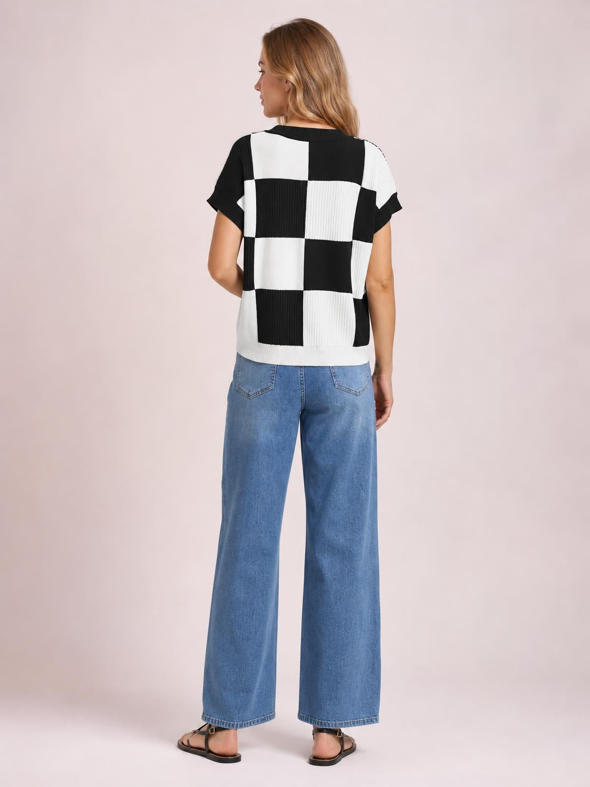 A standing model shows the back of a black-and-white checkered knit sweater with a round neck, short sleeves, and relaxed straight silhouette.