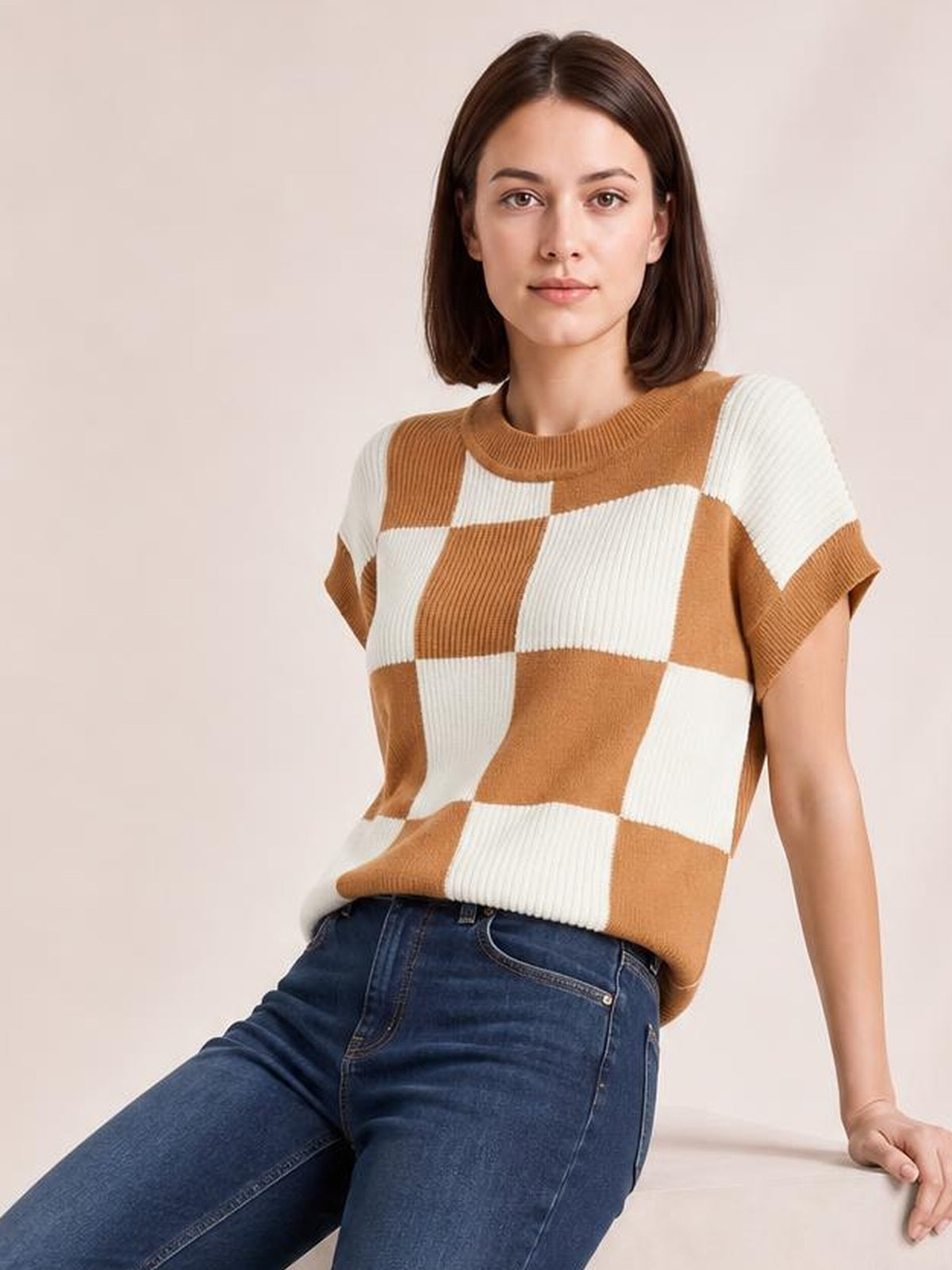 A model sits leaning back in a tan and ivory checkered sweater with a ribbed round neck, short sleeves, and a relaxed silhouette.