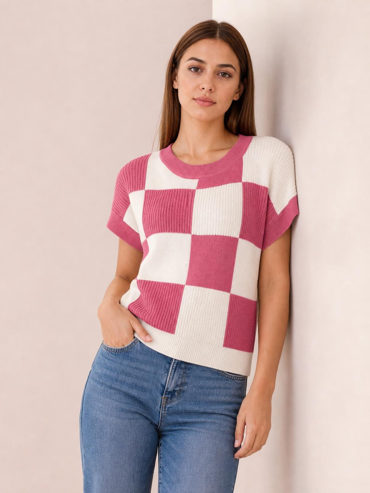 Model wearing a pink and cream checkered round neck short sleeve sweater with ribbed trim and a relaxed silhouette, posing with one hand in her jeans pocket.