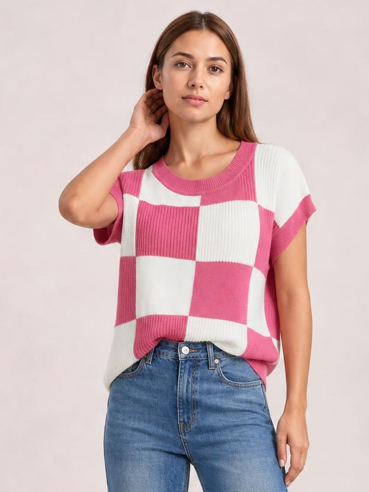 Model wearing a pink and white checkered round-neck short-sleeve sweater with ribbed trim and a relaxed silhouette, posing with one hand at her hair.