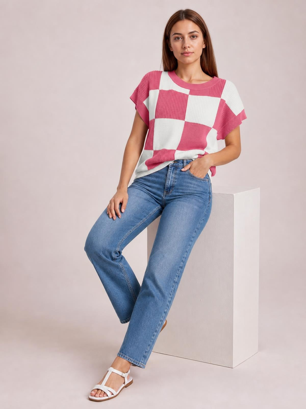 A woman sits with one hand in her jeans pocket, wearing a pink-and-white checkered round-neck short-sleeve sweater with a relaxed silhouette and ribbed knit texture.