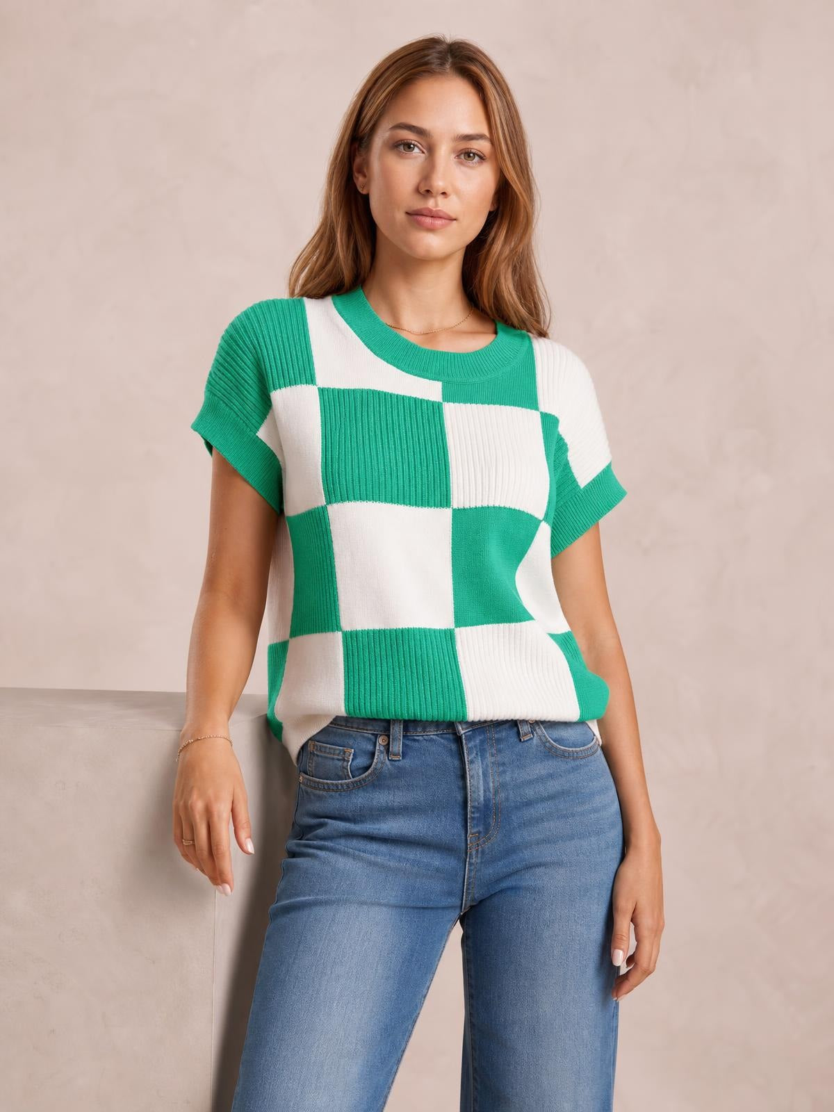 Model wears a green and white checkered round-neck short-sleeve sweater with ribbed trim and a relaxed straight silhouette, posing beside a ledge.