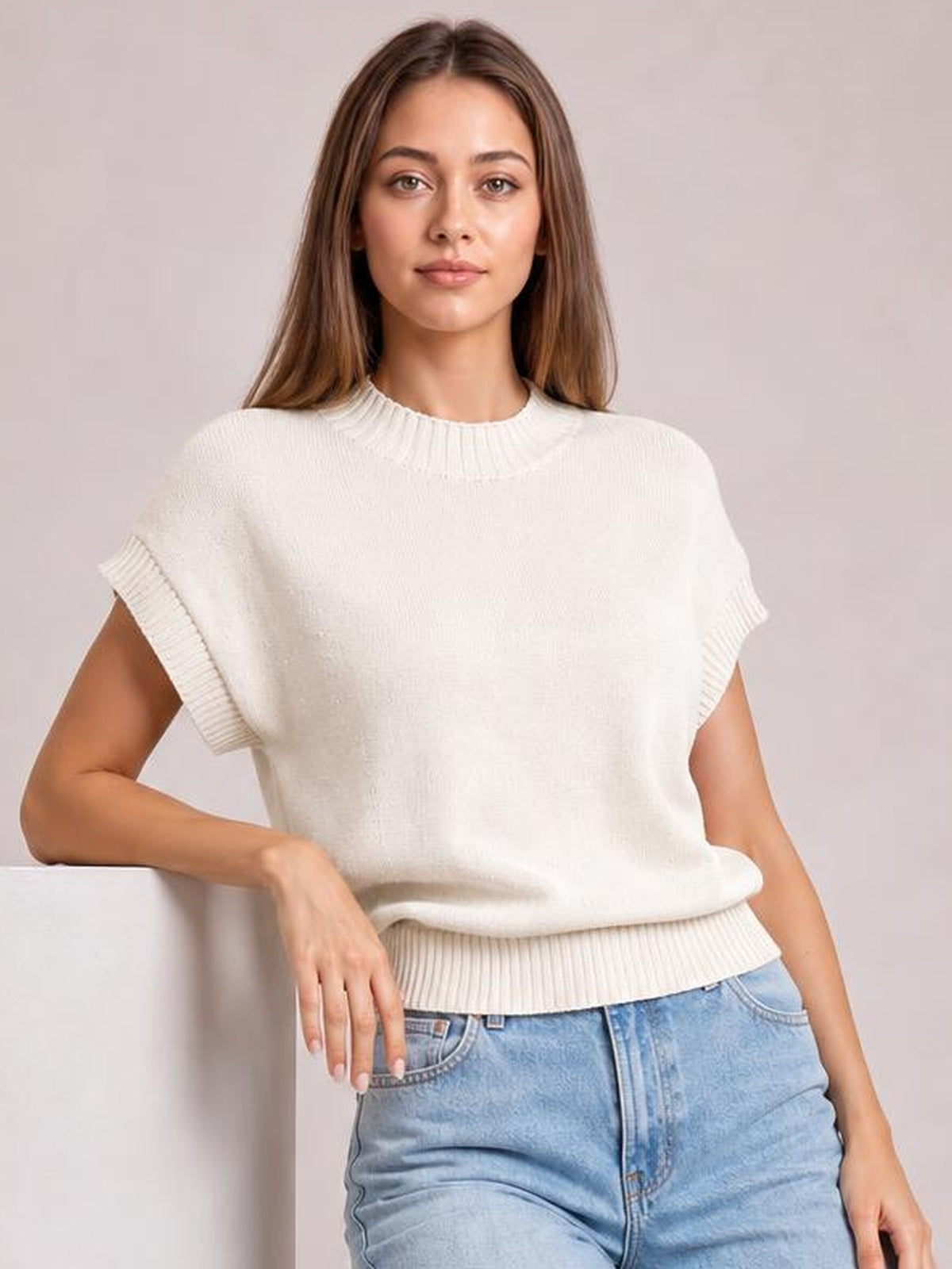 A model wears a cream mock neck short sleeve sweater with a relaxed boxy silhouette, ribbed neckline, cuffs and hem, posing with one hand resting at her side.