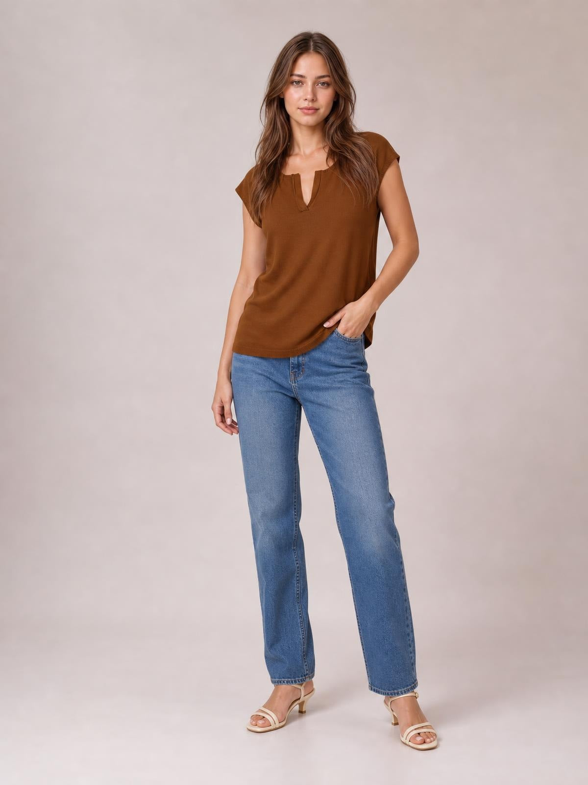 Model wears a brown notched cap sleeve knit top with a relaxed straight silhouette and ribbed hem, paired with jeans while standing with one hand in a pocket.