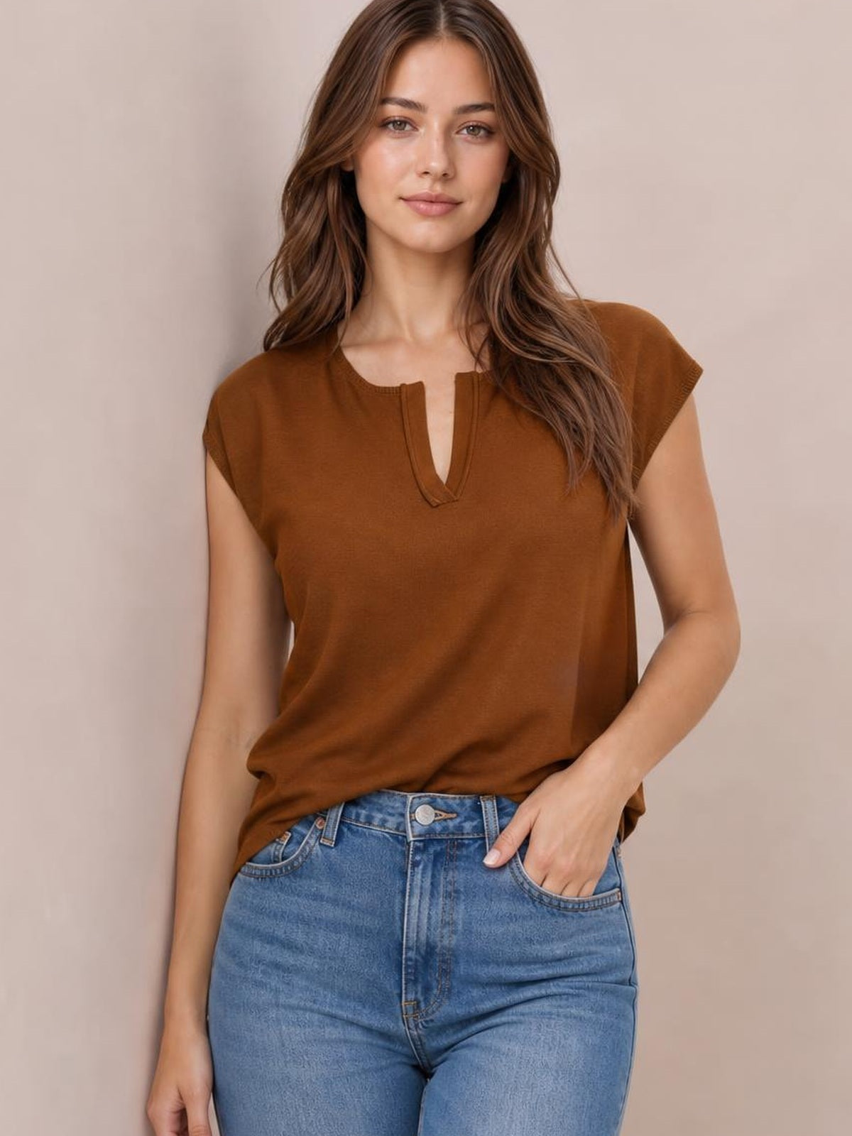 Model wearing a brown notched cap sleeve knit top with a relaxed silhouette and visible V-shaped neckline detail, one hand in her jeans pocket.