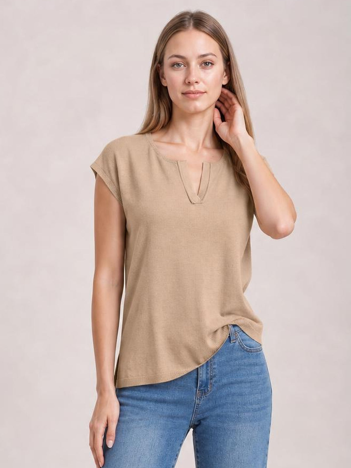 Model poses in a beige knit top with notched neckline, cap sleeves, straight relaxed silhouette, and ribbed trim, paired with blue jeans.