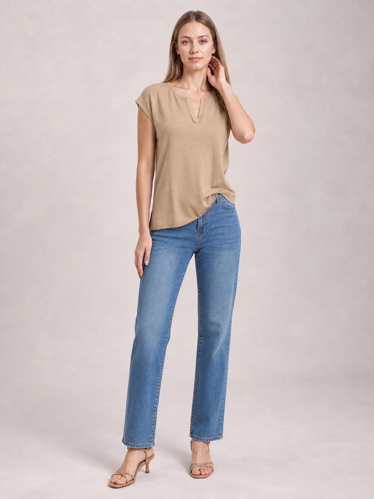 Model wears a beige notched cap sleeve knit top with a relaxed straight silhouette, standing in jeans with one hand resting near her neck.