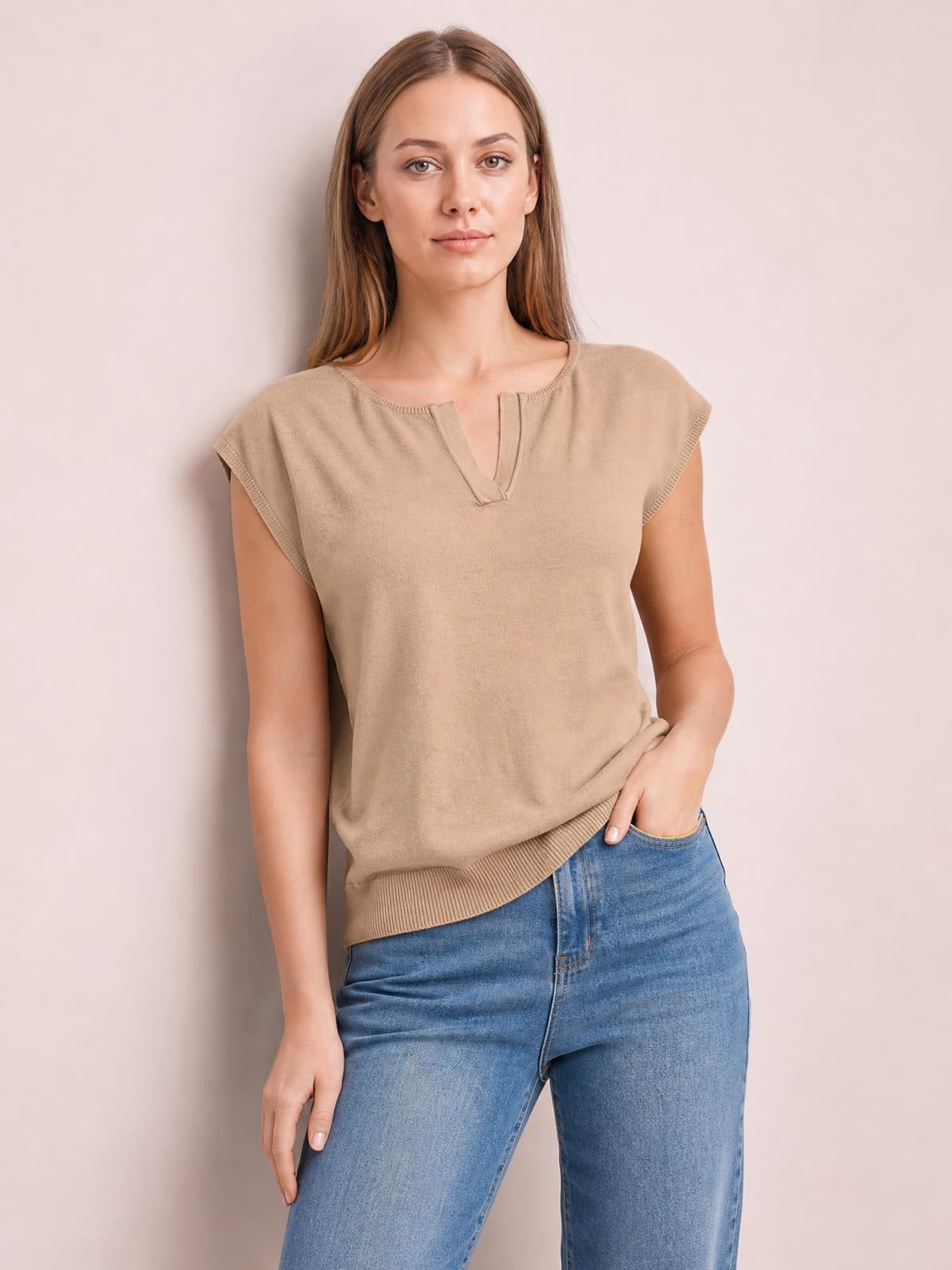 Model poses in a beige notched cap sleeve knit top with a relaxed silhouette, ribbed neckline and hem, and slight stretch, paired with jeans.