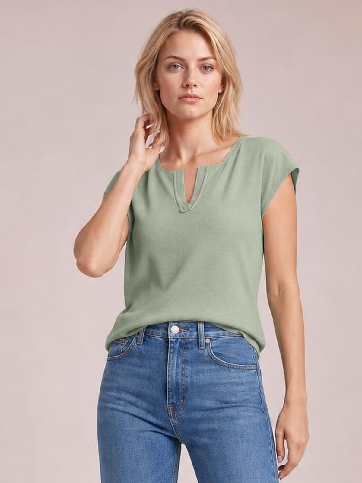 Model poses in a sage green notched cap sleeve knit top with a relaxed straight silhouette and subtle neckline trim, paired with blue jeans.