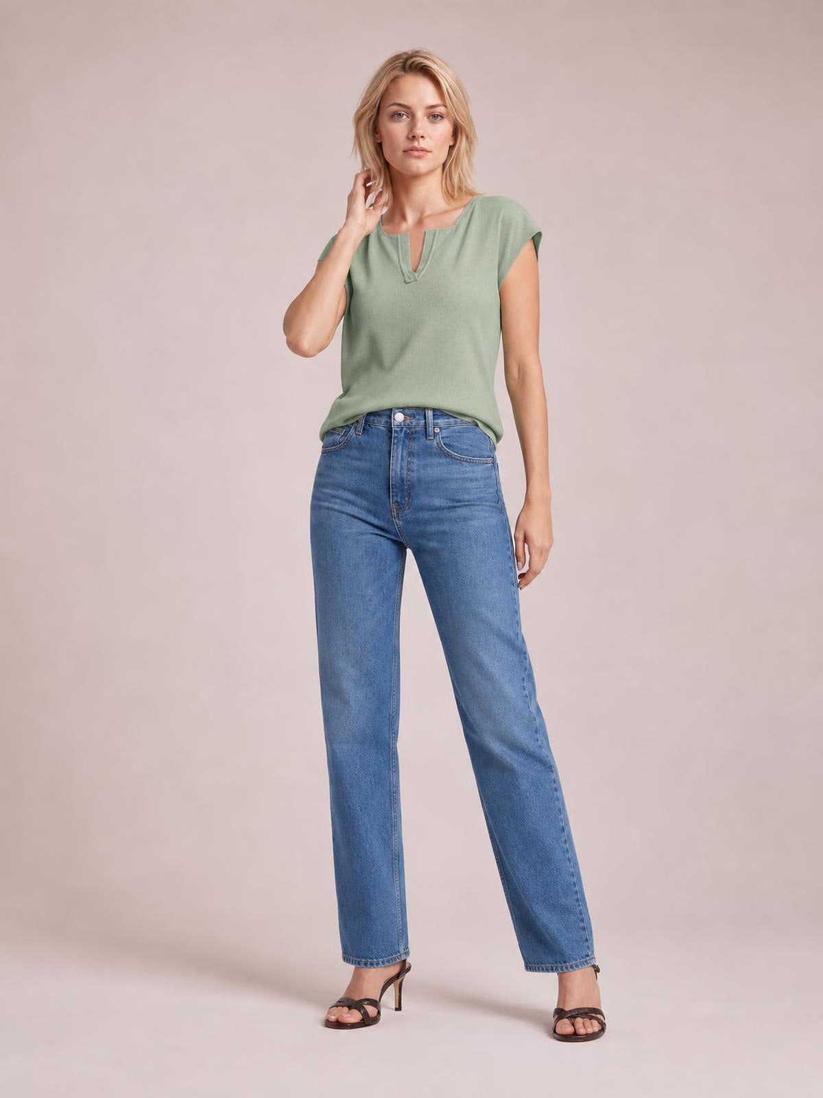 Model poses in a sage green notched cap sleeve knit top with a relaxed straight silhouette and subtle neckline slit, paired with blue jeans.