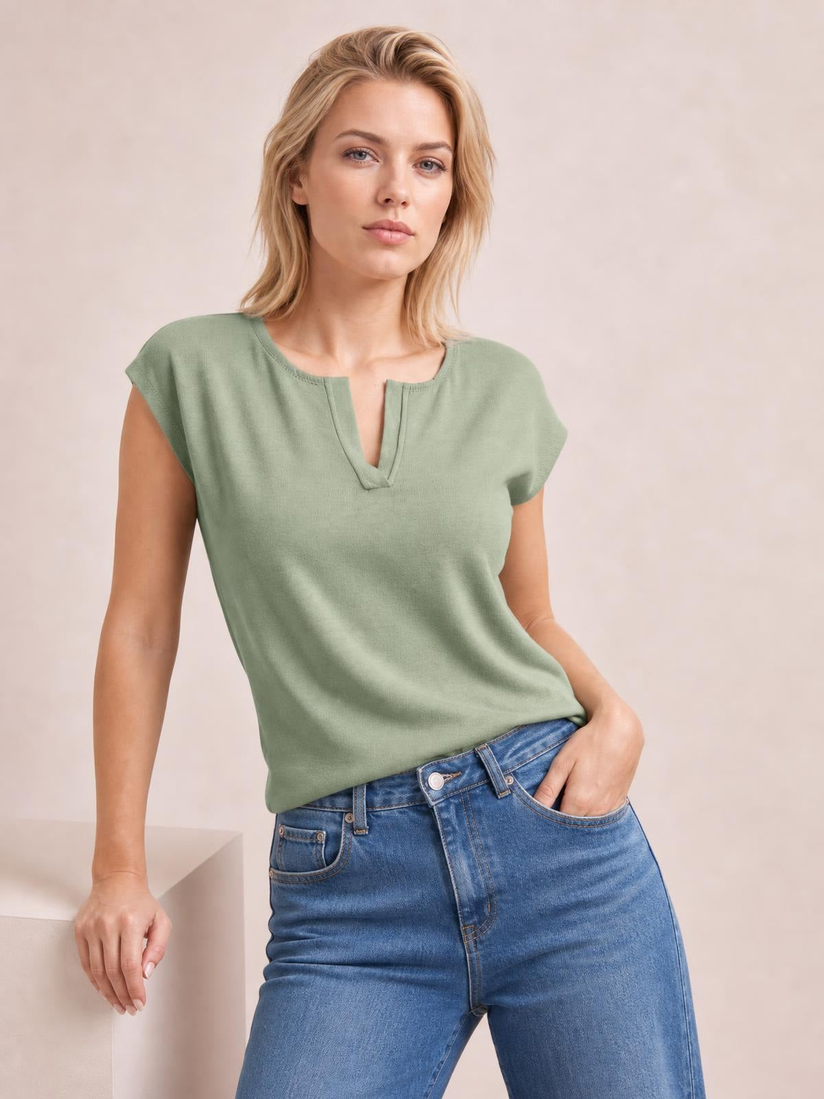 Model wearing a sage green notched cap sleeve knit top with a relaxed straight silhouette, standing with one hand in her jeans pocket.