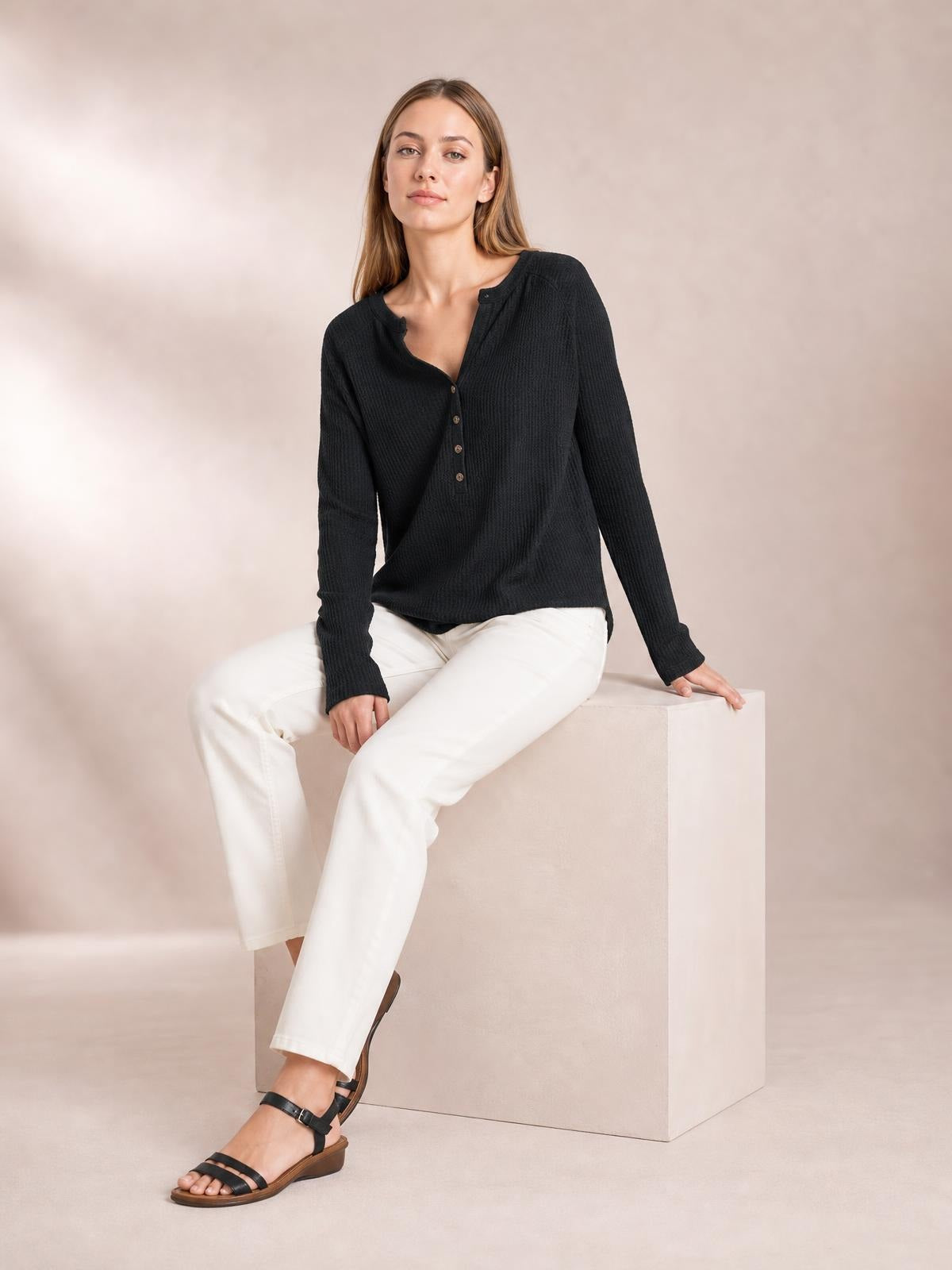 A model sits wearing a black notched thumbhole long sleeve T-shirt with a buttoned neckline, ribbed texture, and a relaxed straight silhouette.