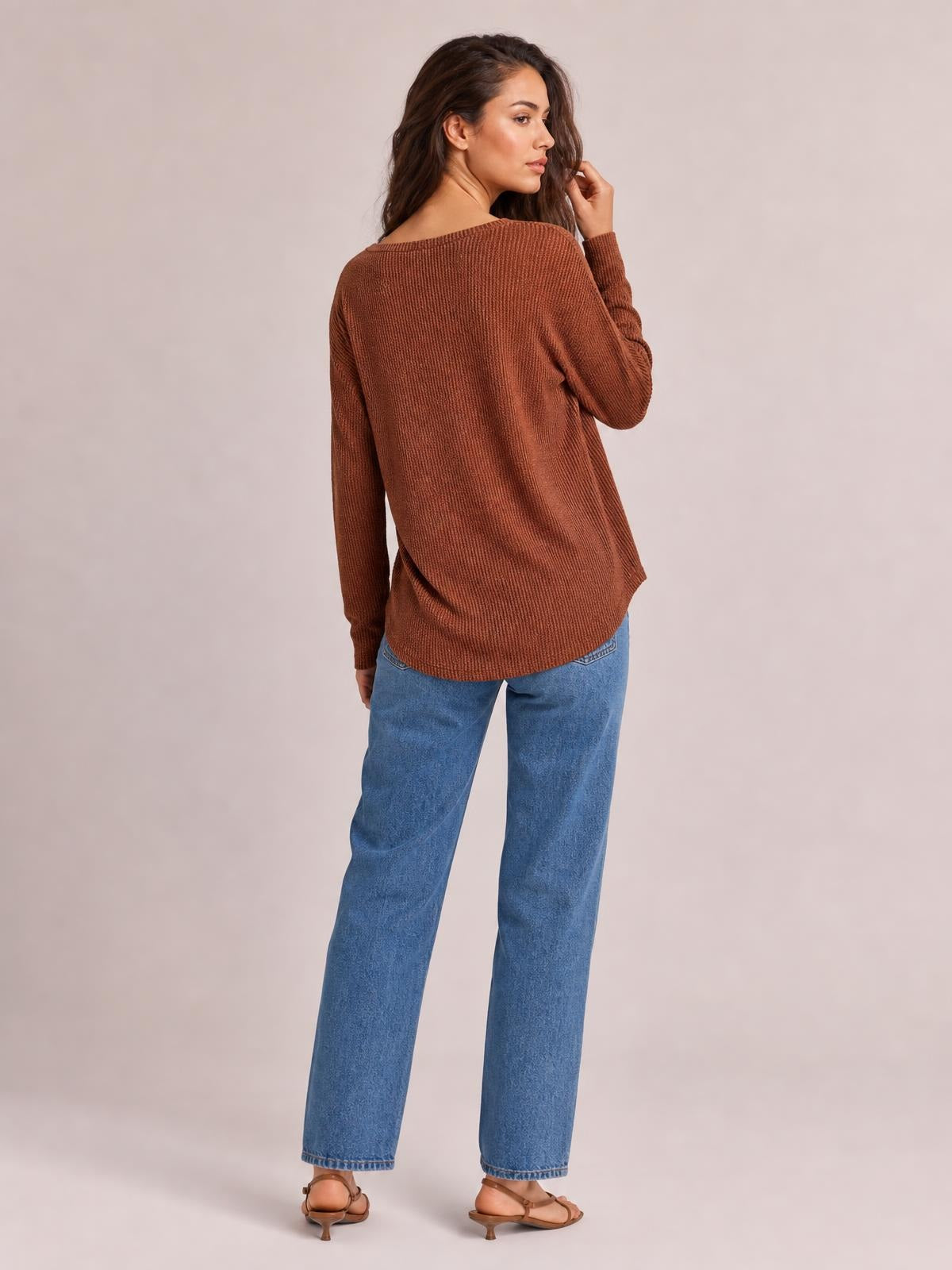 A model poses turned away in a rust-brown ribbed long sleeve T-shirt with a relaxed silhouette, curved hem, and moderate stretch.
