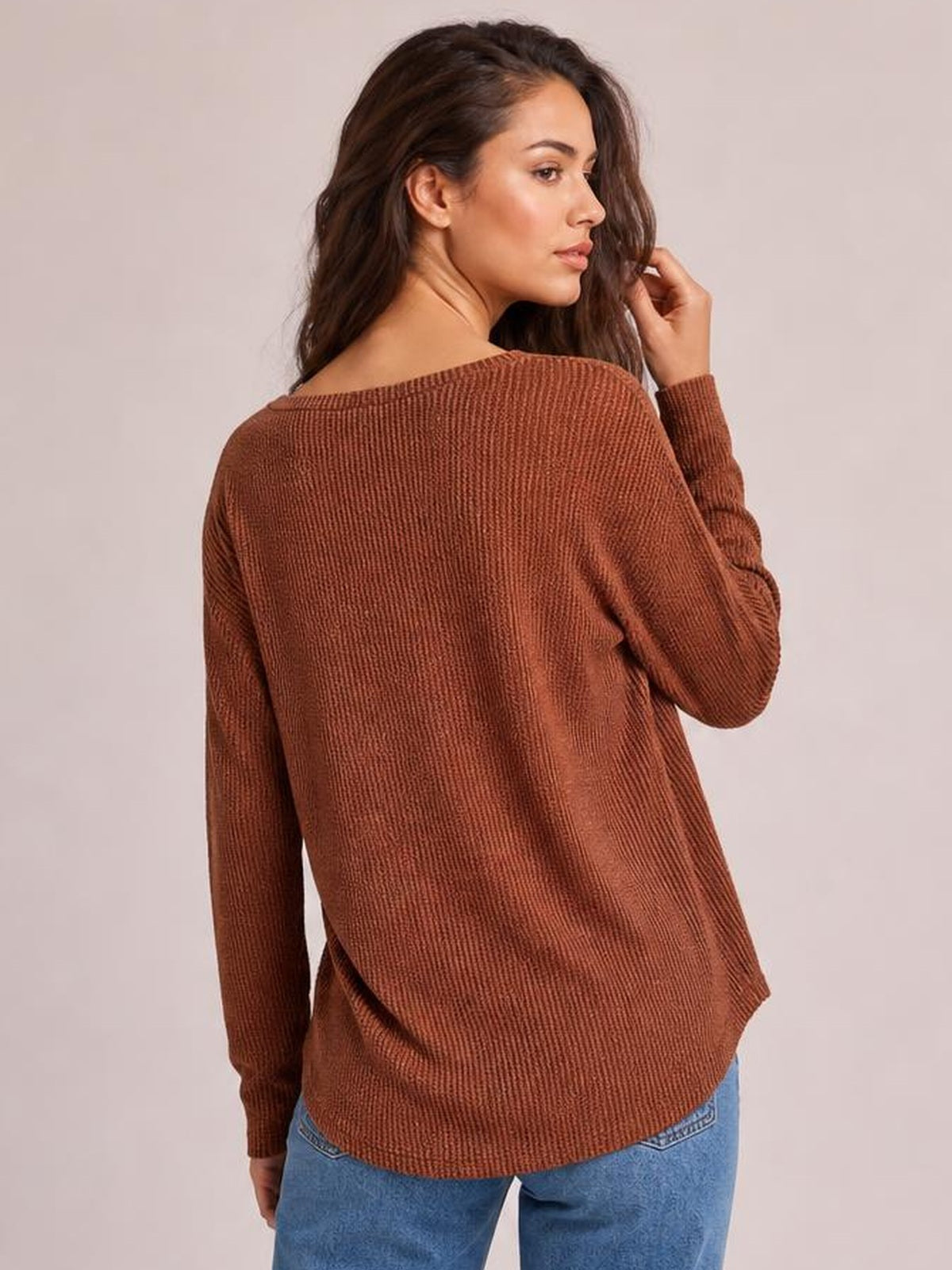 A model poses in a rust-brown long sleeve T-shirt, showing its relaxed silhouette, textured knit construction, curved hem, and wide neckline from the back.