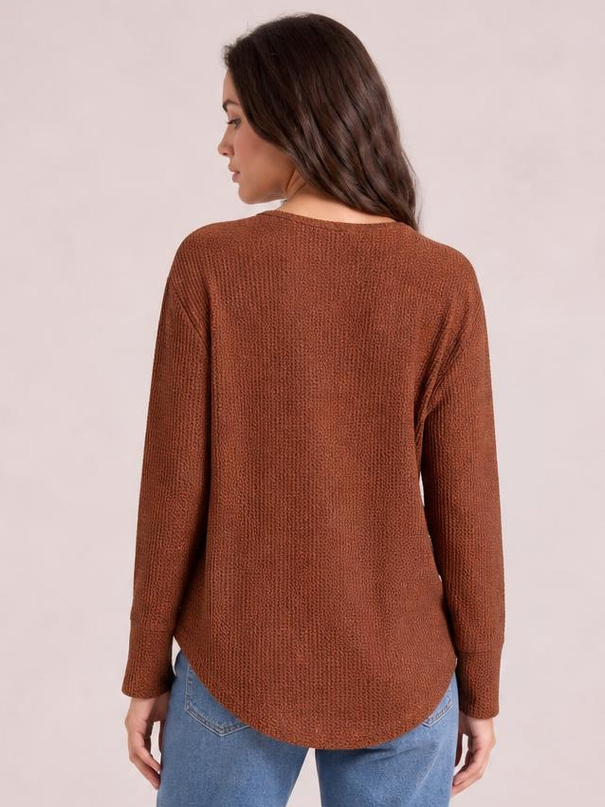 A model looks over her shoulder wearing a rust-brown long sleeve T-shirt with a relaxed silhouette, textured knit construction, and rounded hem over jeans.