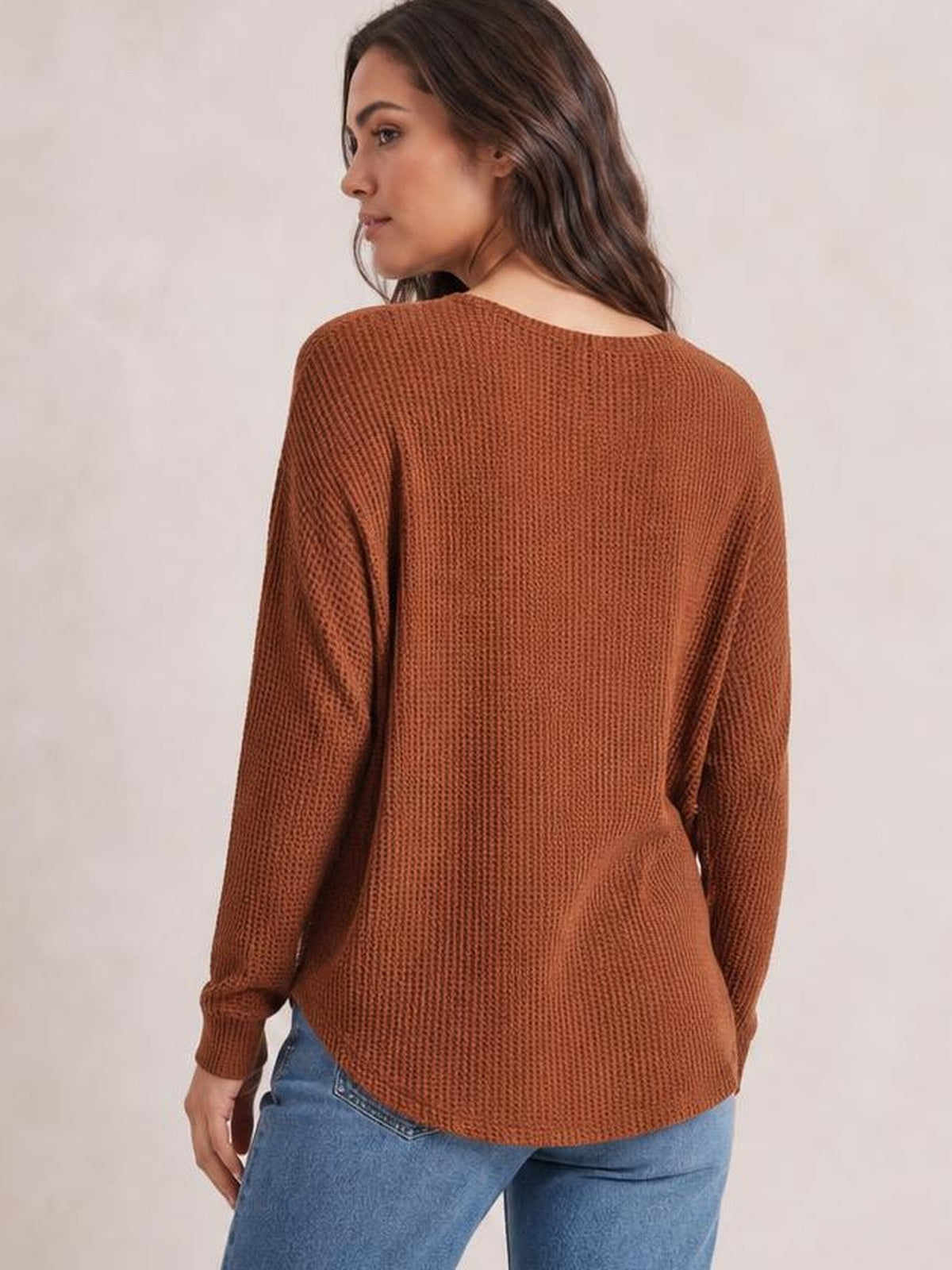 A model faces away wearing a rust-brown long-sleeve T-shirt with textured knit construction, a relaxed silhouette, fitted cuffs, and a curved hem.