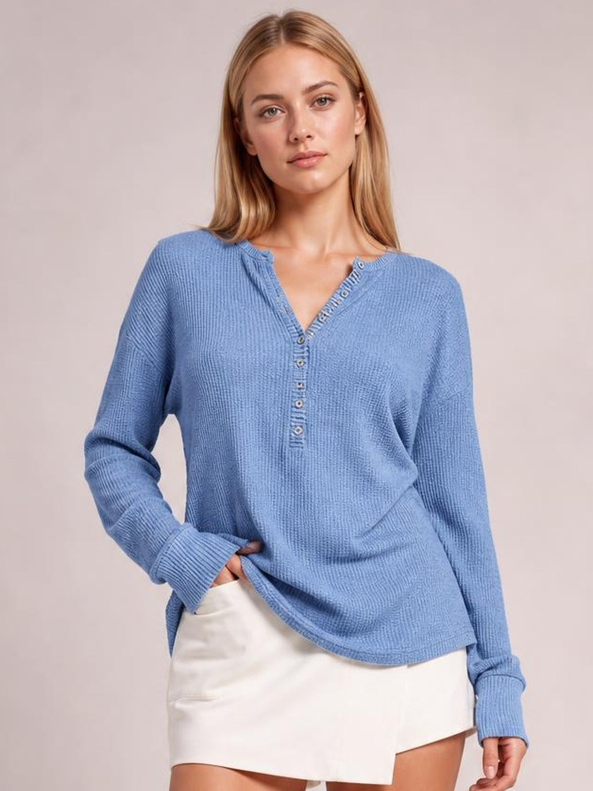 Model wears a light blue notched long sleeve T-shirt with button details, extended cuffs, a relaxed silhouette, and a curved hem while posing with one hand at the side.