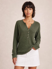 Model wearing an olive green notched long sleeve T-shirt with a buttoned front, ribbed texture, relaxed silhouette, and one hand tucked into a pocket.