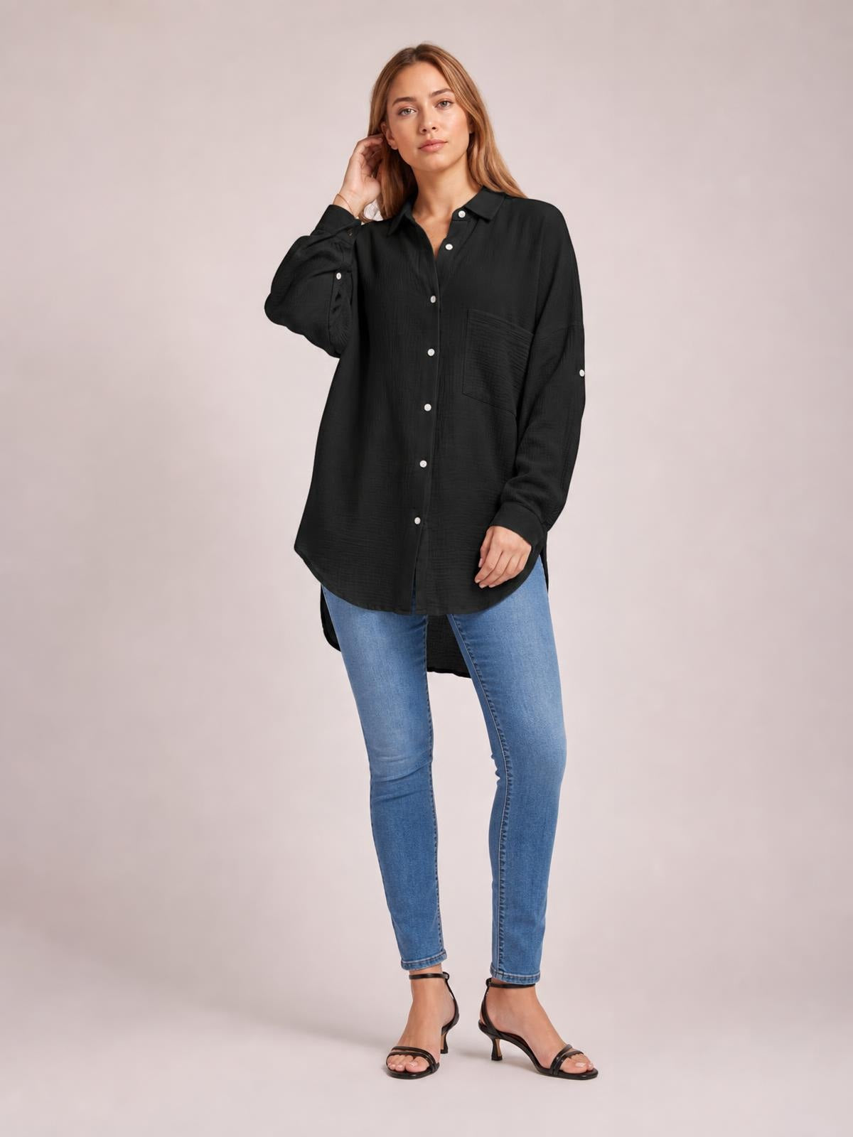 A model poses in a black textured button-up shirt with long sleeves, a relaxed silhouette, chest pocket, white buttons, and a curved hem.