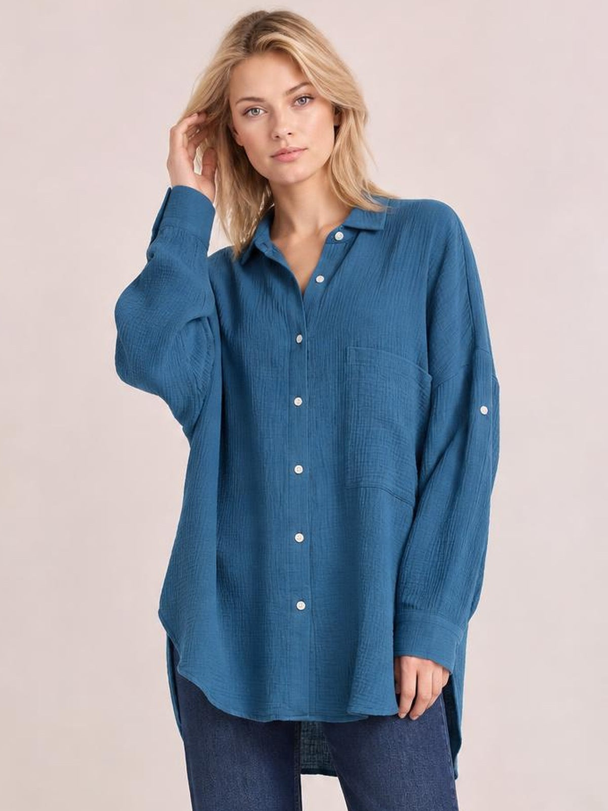 Model wears a blue textured button-up shirt with a pointed collar, long sleeves, chest pocket, curved hem, and relaxed tunic silhouette while touching her hair.