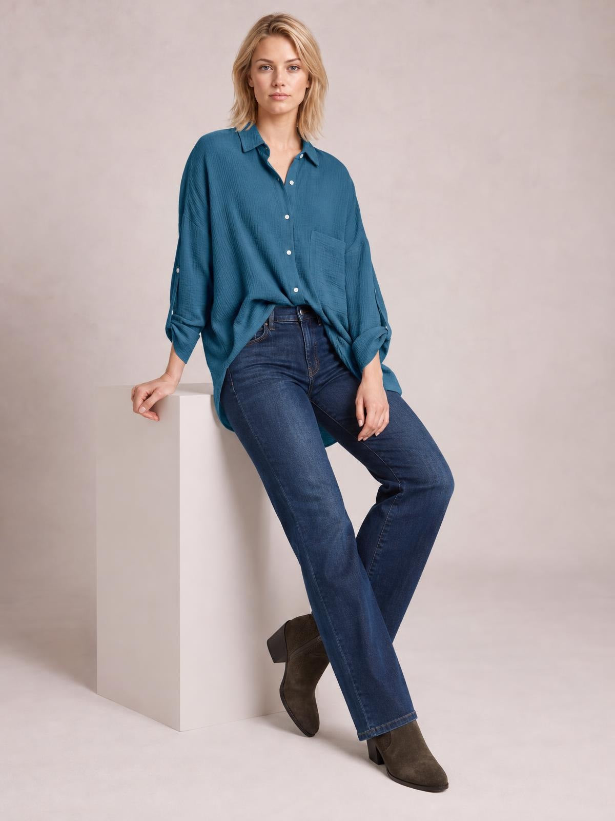 Model sits with crossed legs wearing a teal textured button-up shirt with a relaxed silhouette, front buttons, chest pocket, and rolled sleeves.
