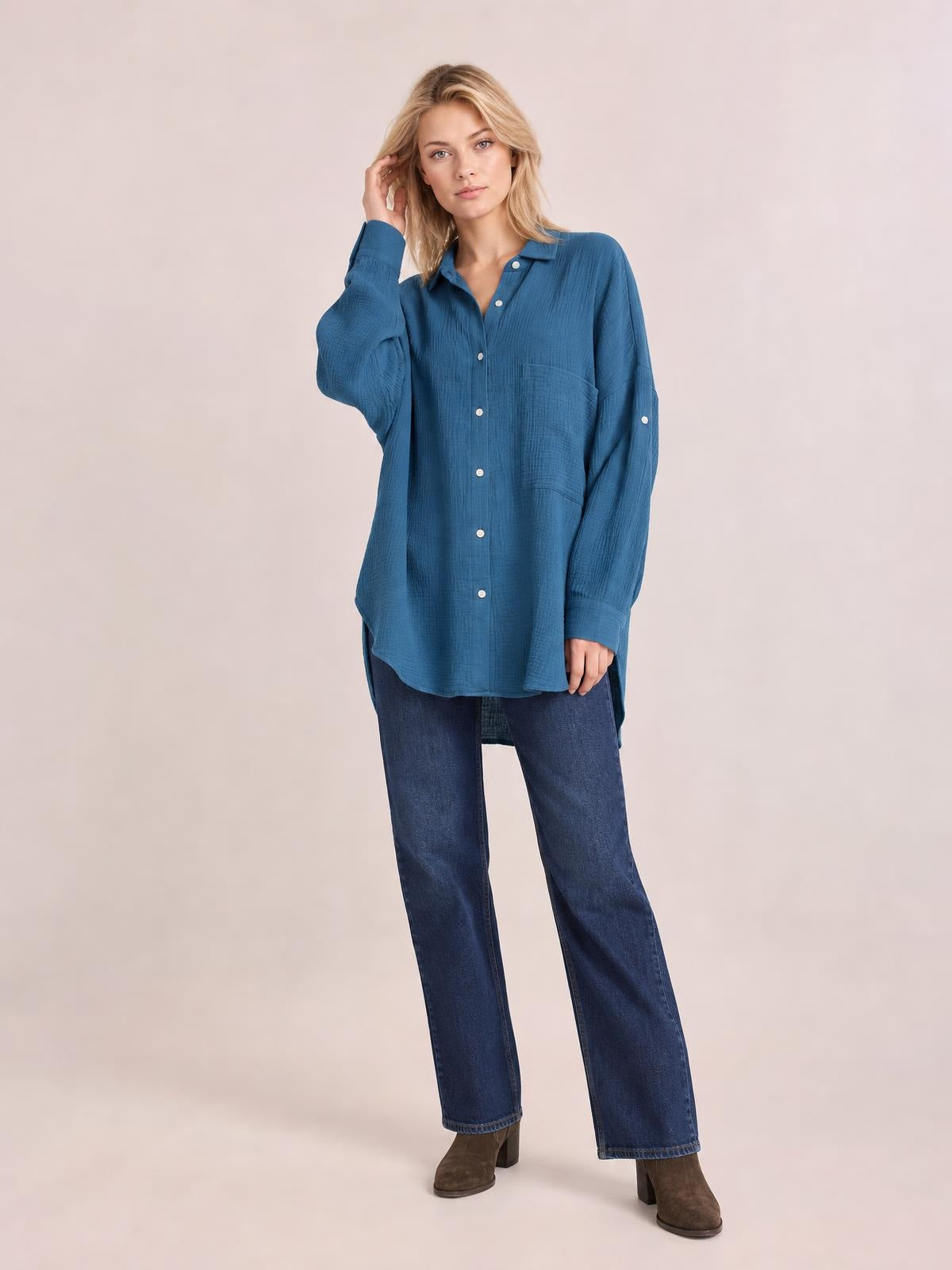 Model poses in a textured blue button-up shirt with long sleeves, a collared neckline, front buttons, chest pocket, and relaxed longline silhouette.