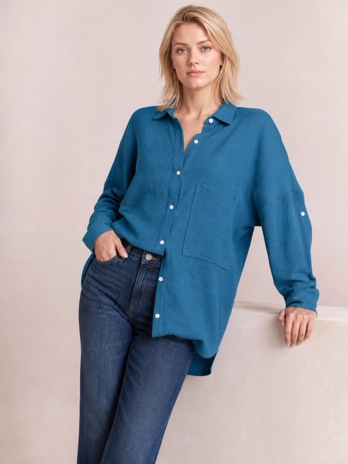 A model poses in a relaxed teal blue textured button-up shirt with a collared neckline, white buttons, long sleeves, and a loose pocketed silhouette.