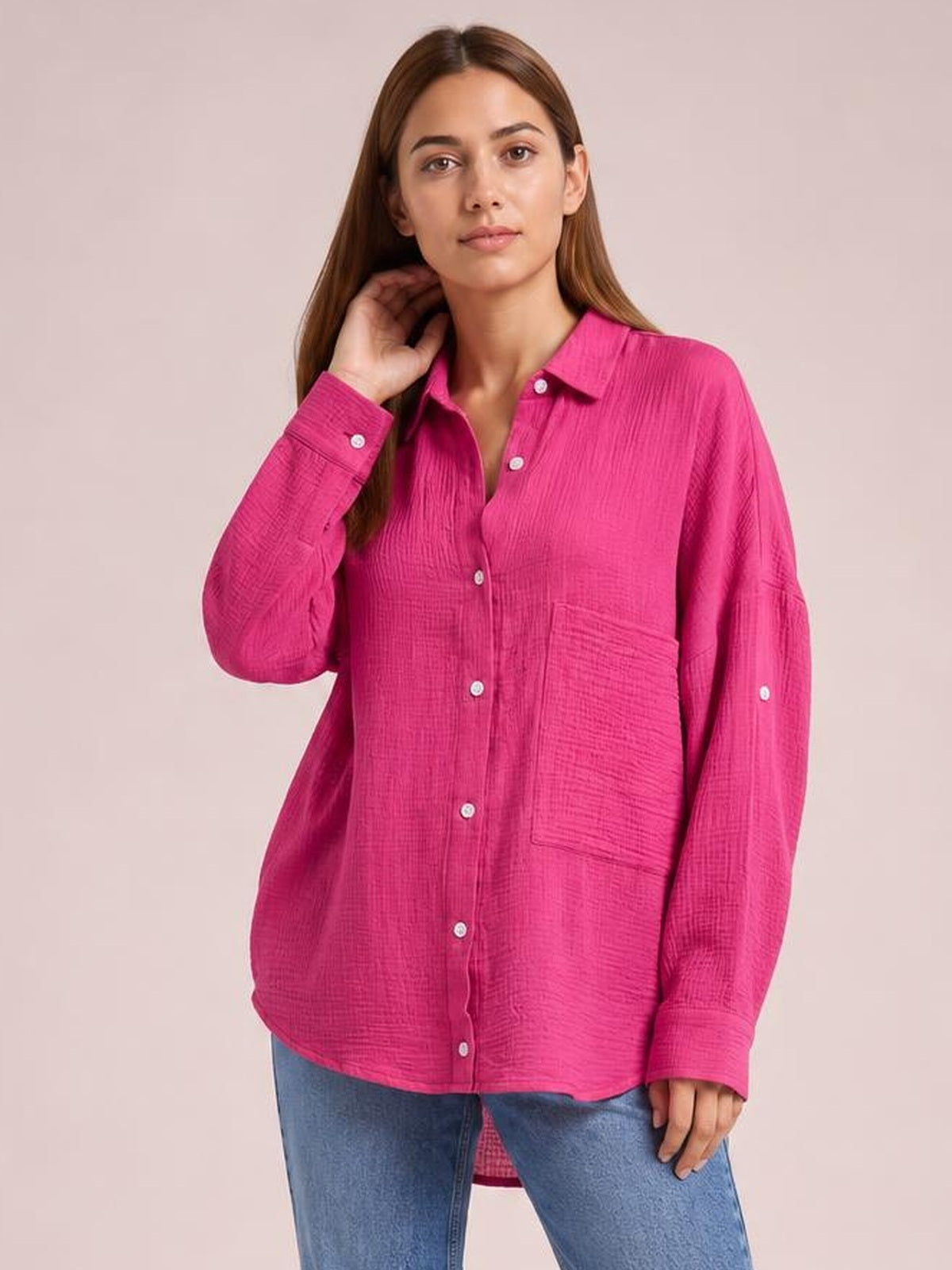 A model touches her hair while wearing a bright pink textured button-up shirt with a collar, long sleeves, relaxed silhouette, and visible chest pockets.