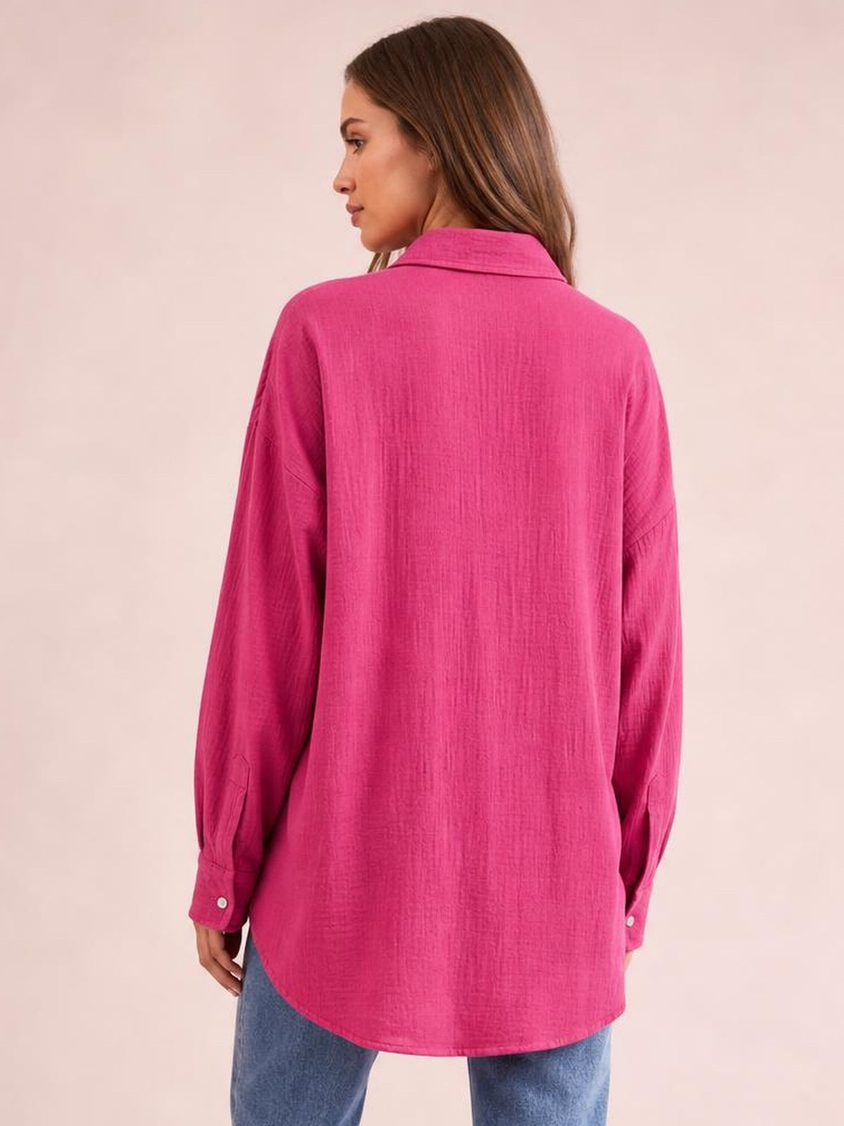 A model shows the back of a bright pink textured button-up shirt with a collar, long sleeves, relaxed oversized silhouette, and curved hem.