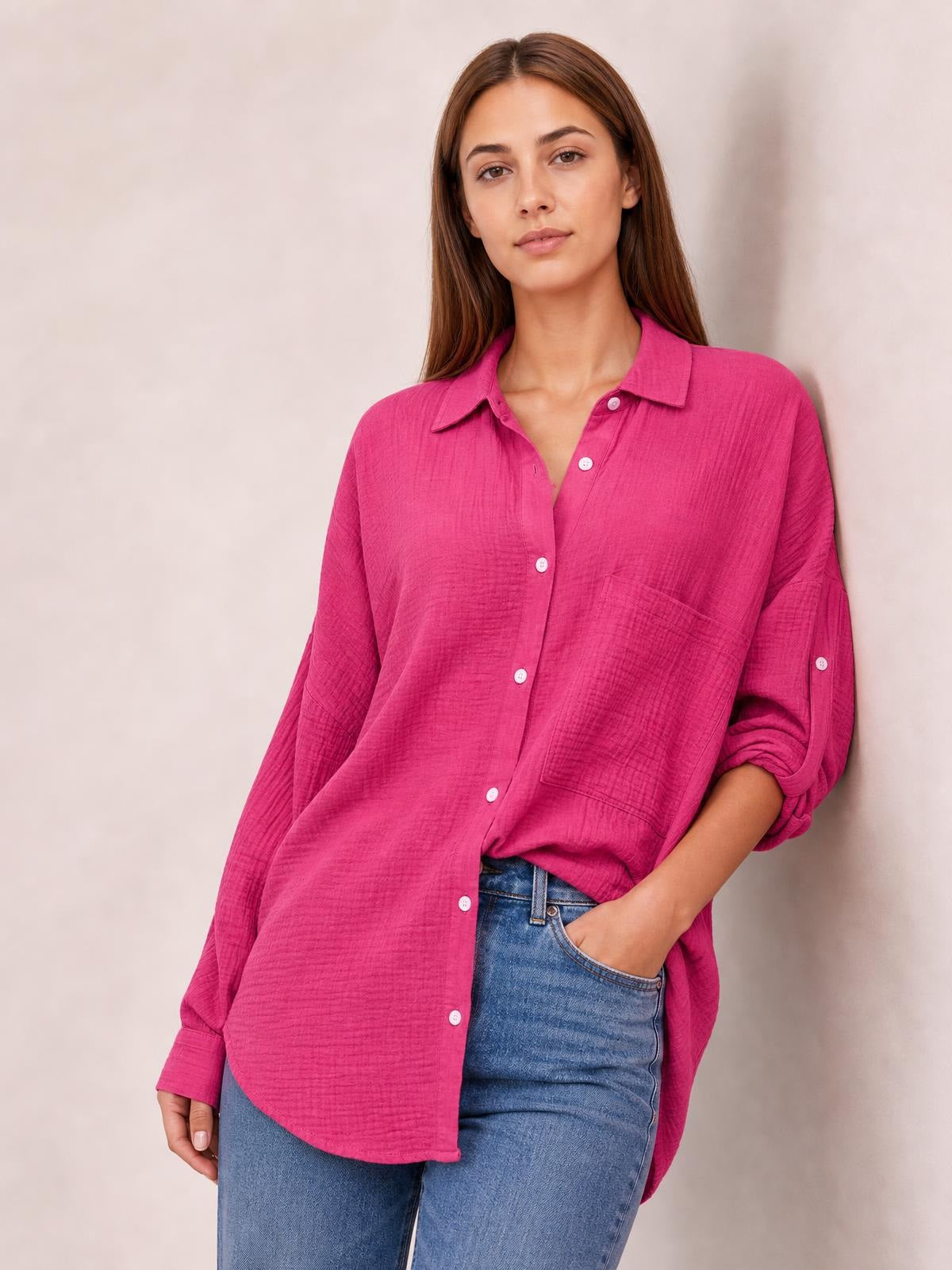 Model wears a bright pink textured button-up shirt with a relaxed silhouette, pointed collar, chest pocket, curved hem, and rolled sleeves while posing with one hand in her jeans pocket.