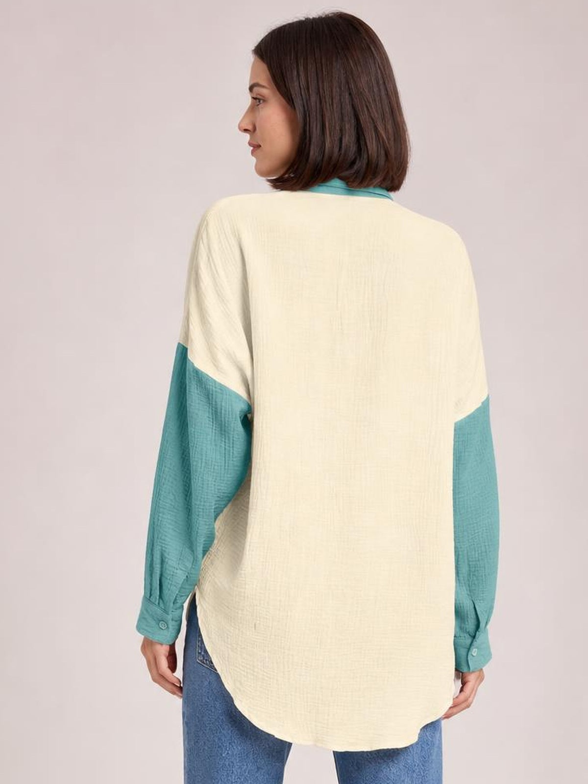 A model stands turned away in a cream textured button-up shirt with contrasting teal long sleeves and a relaxed, rounded-hem silhouette.