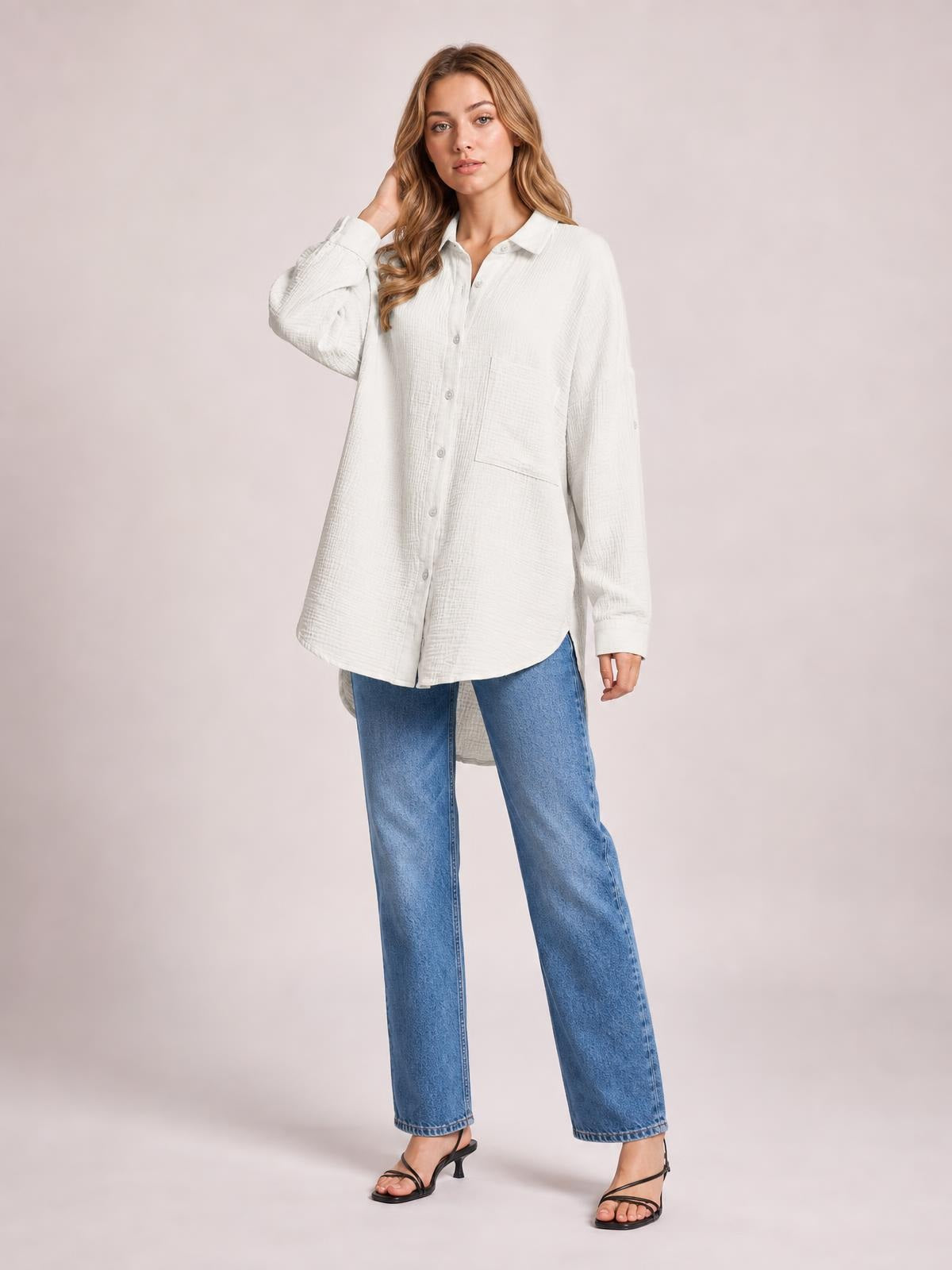 A model poses with one hand in her hair, wearing a white textured button-up shirt with a collar, long sleeves, front buttons, a pocket, and a relaxed longline silhouette.