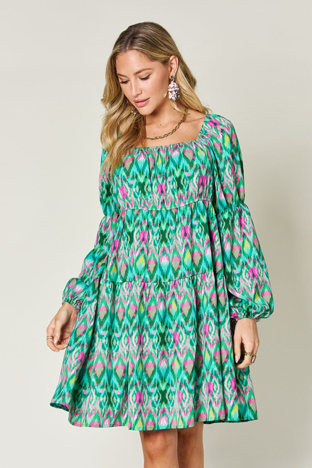Model wears a vibrant green and pink printed long sleeve dress with a square neckline, gathered waist, tiered A-line silhouette, and balloon sleeves.