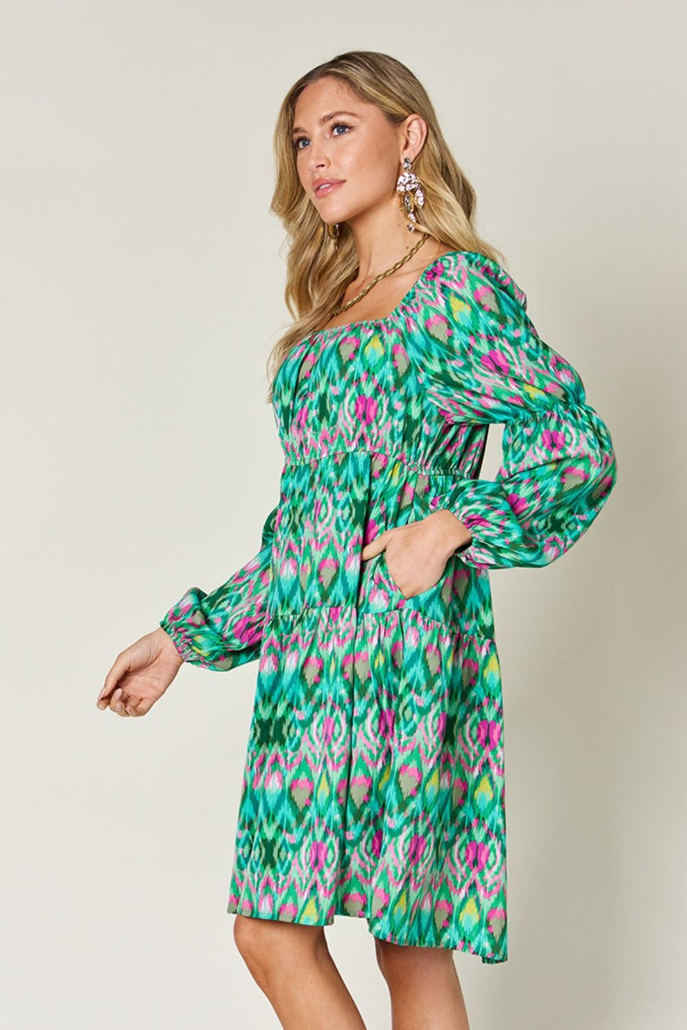 Model poses in a vibrant green printed long sleeve dress with pink and yellow accents, a gathered waist, tied neckline, balloon sleeves, and easy A-line silhouette.